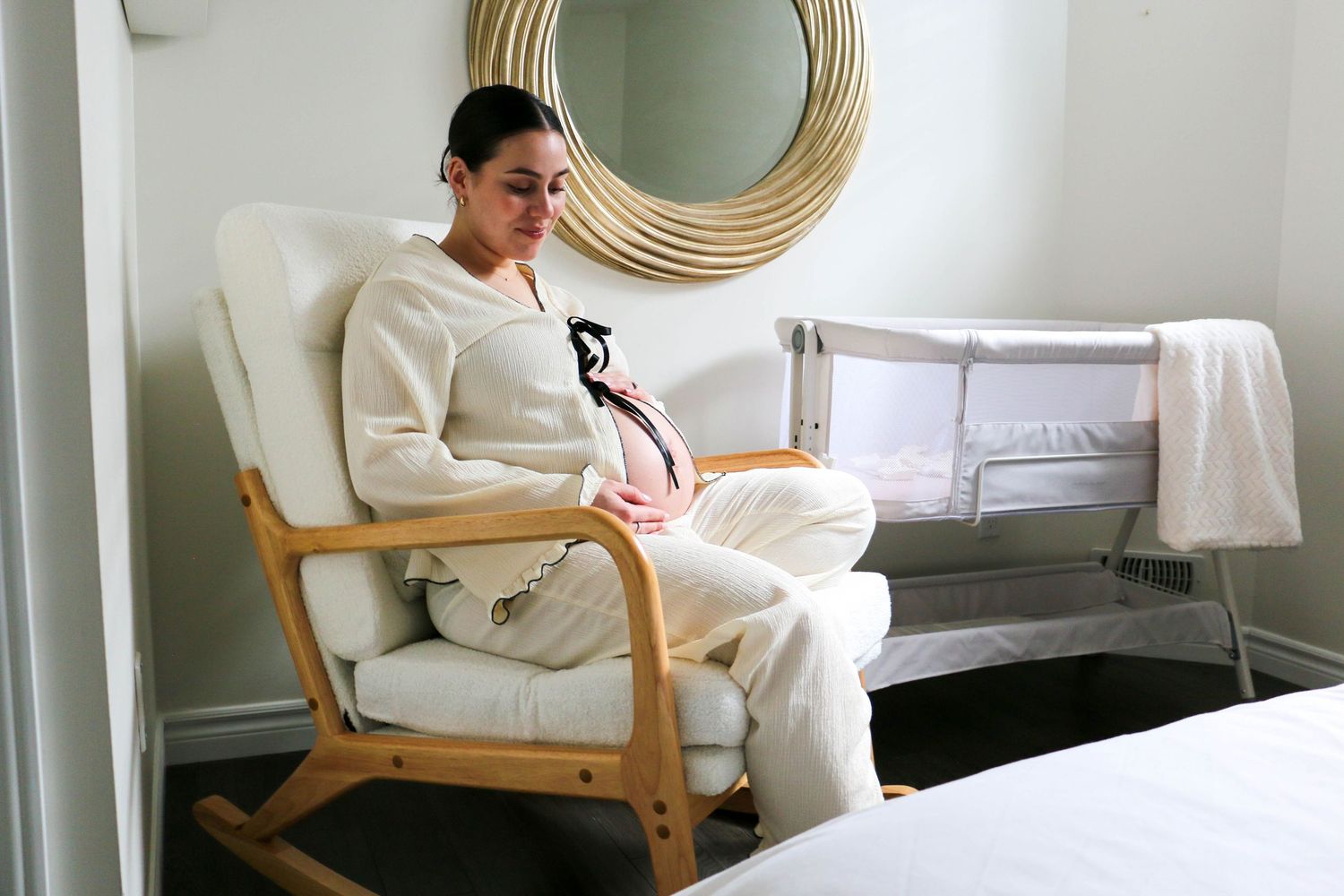 Person in white sweater sits in wooden chair reading near circular mirror and window in bright minimal space.