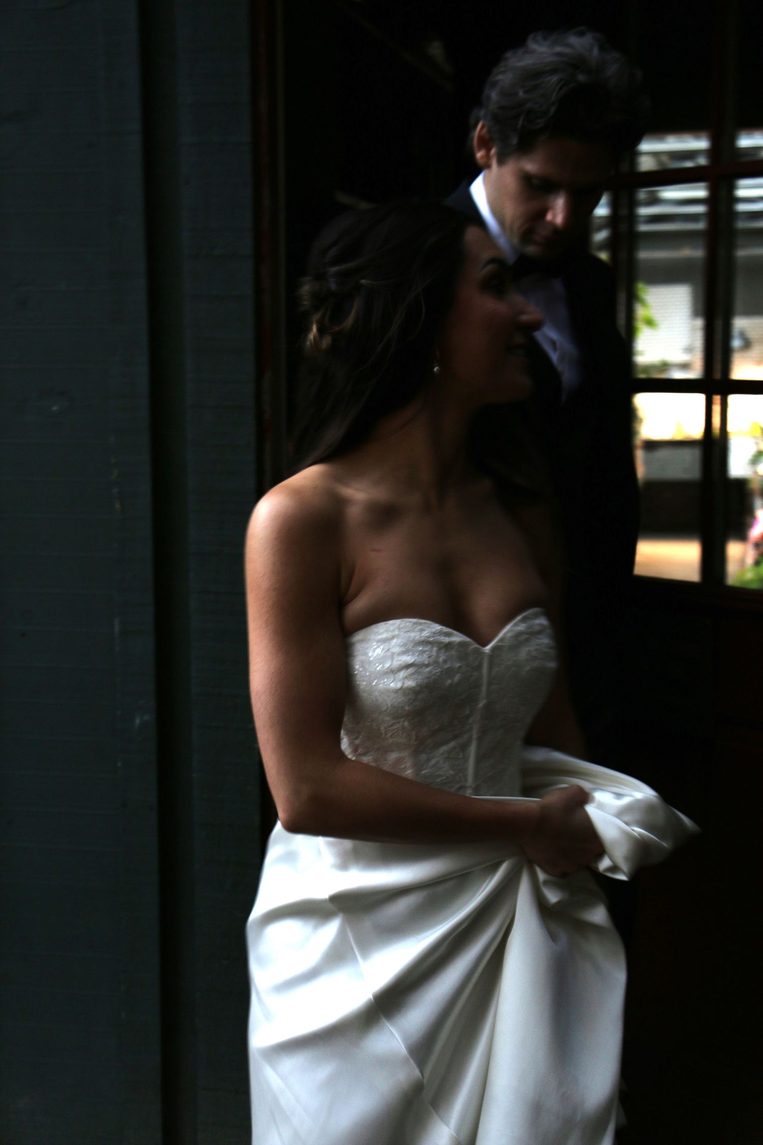 Bride in white dress stands in dramatic lighting near window.