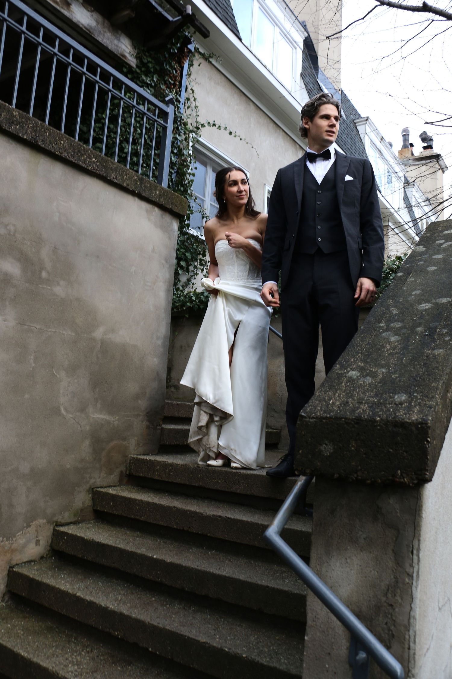Couple in formal wedding attire descending stone steps against urban backdrop.