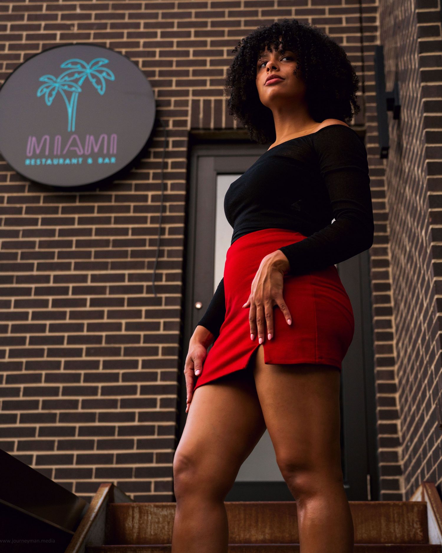 A person wearing black and red attire poses against a brick wall backdrop with a neon palm tree sign.