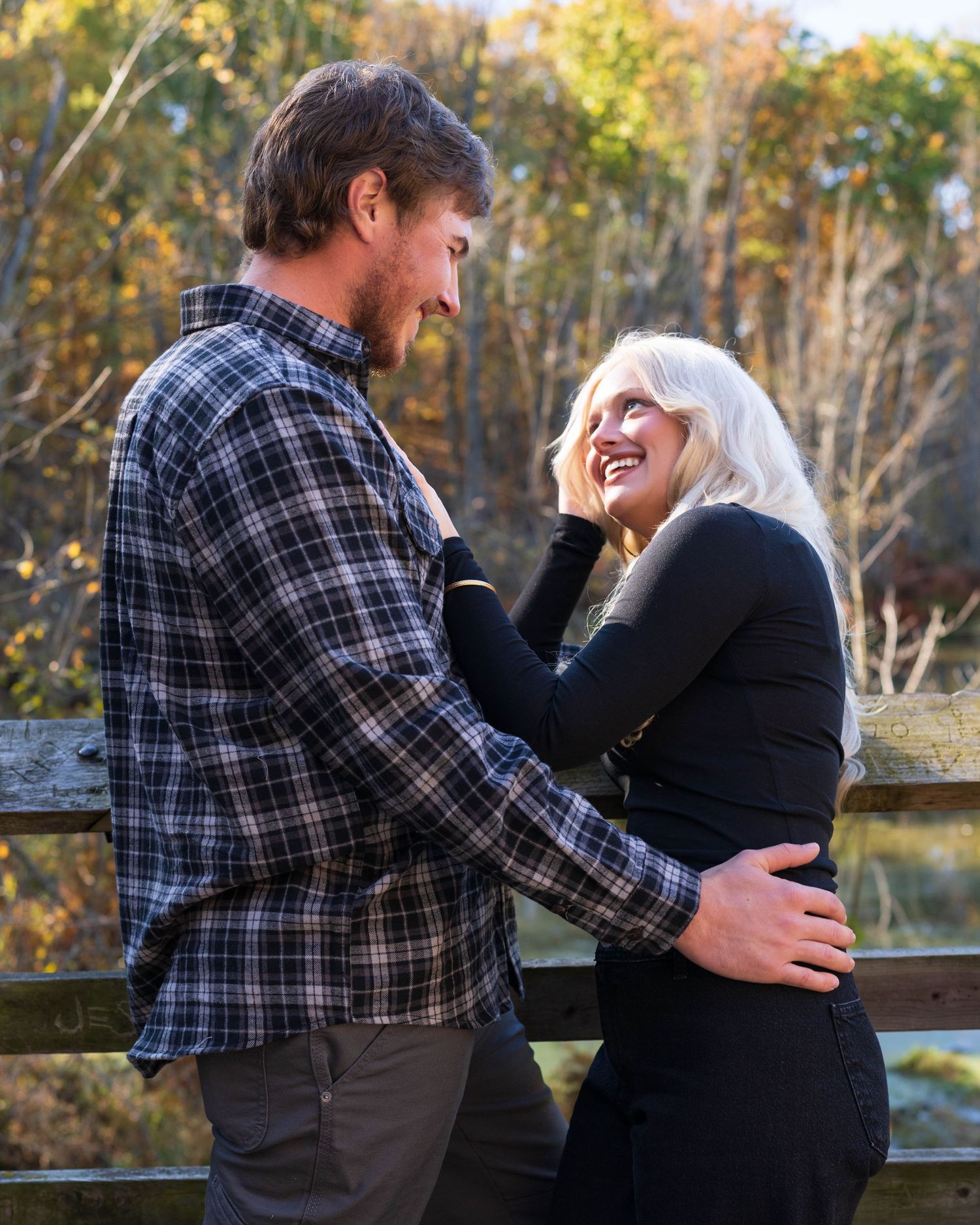 A couple shares a joyful moment on a wooden bridge surrounded by autumn foliage, the woman laughing in a black outfit.