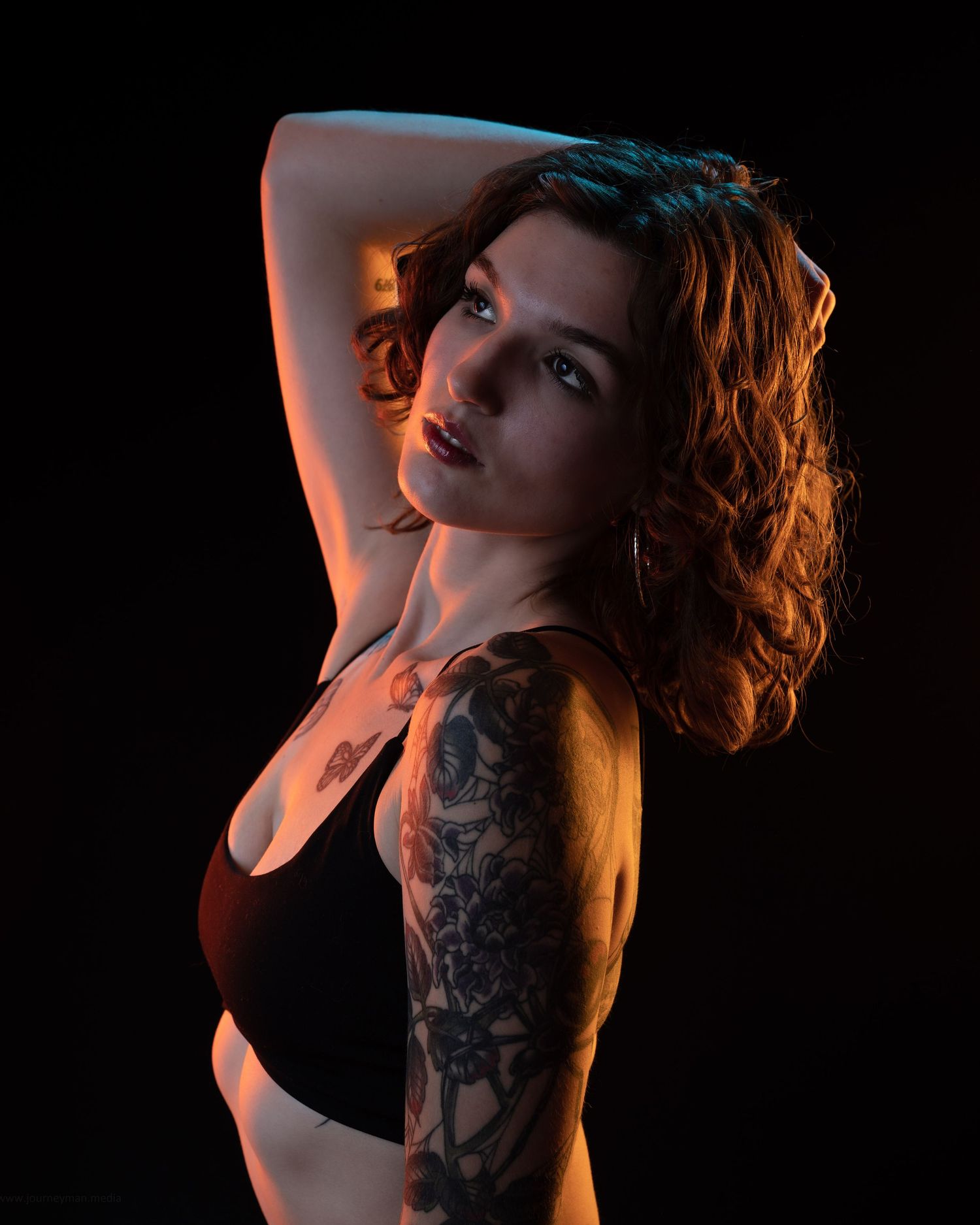 A tattooed redhead woman poses confidently in a black crop top against a dark background with dramatic warm lighting.