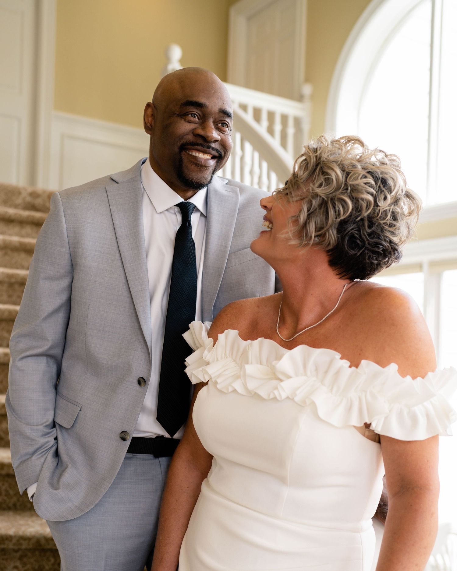 A smiling couple in wedding attire, man in gray suit and woman in white off-shoulder dress, posing on elegant staircase.