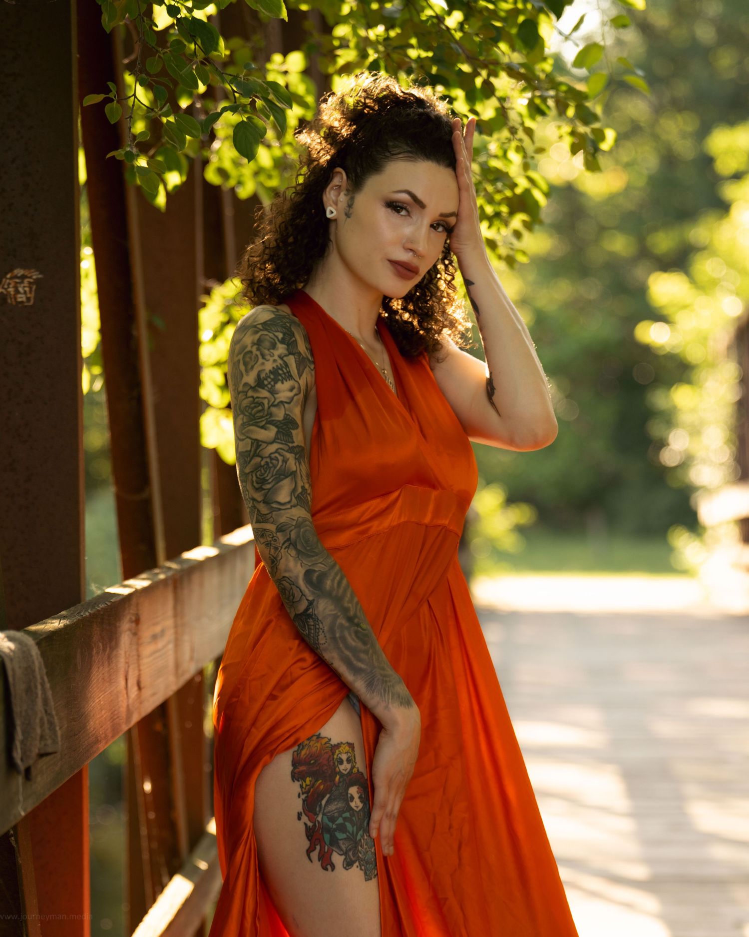 Model in flowing orange dress poses gracefully on wooden bridge with sunlit trees in background.