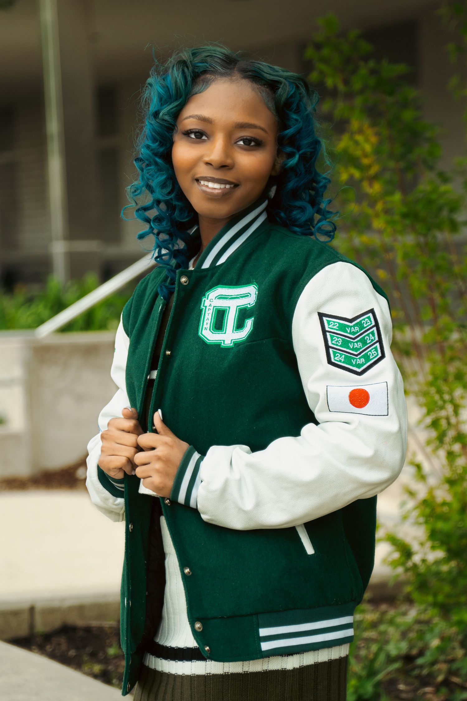 Student wearing a green and white varsity letterman jacket with school logo and patches poses outdoors with a bright smile.