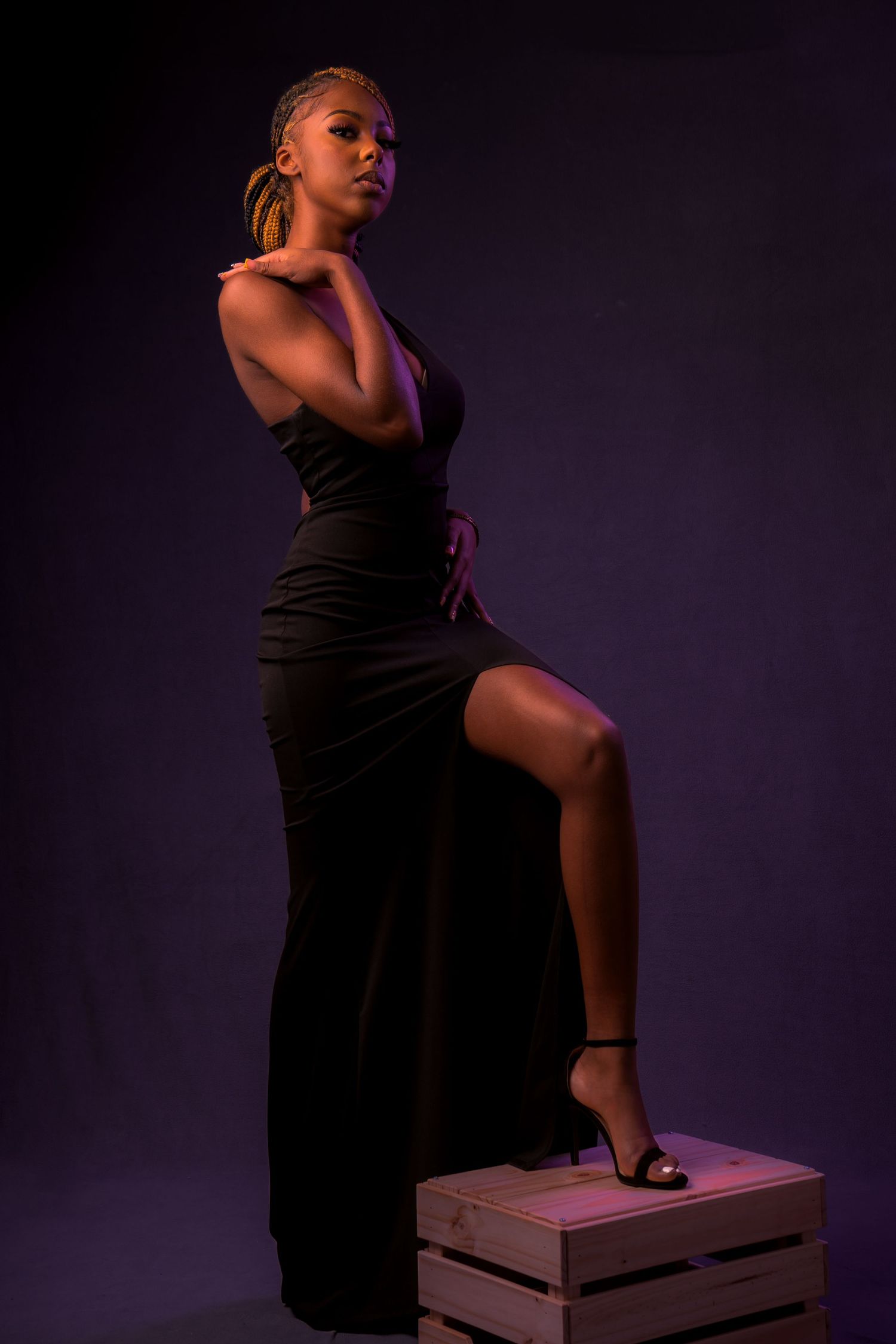 A figure in a black dress poses elegantly on wooden boxes against a dark purple studio backdrop in a series of photos.