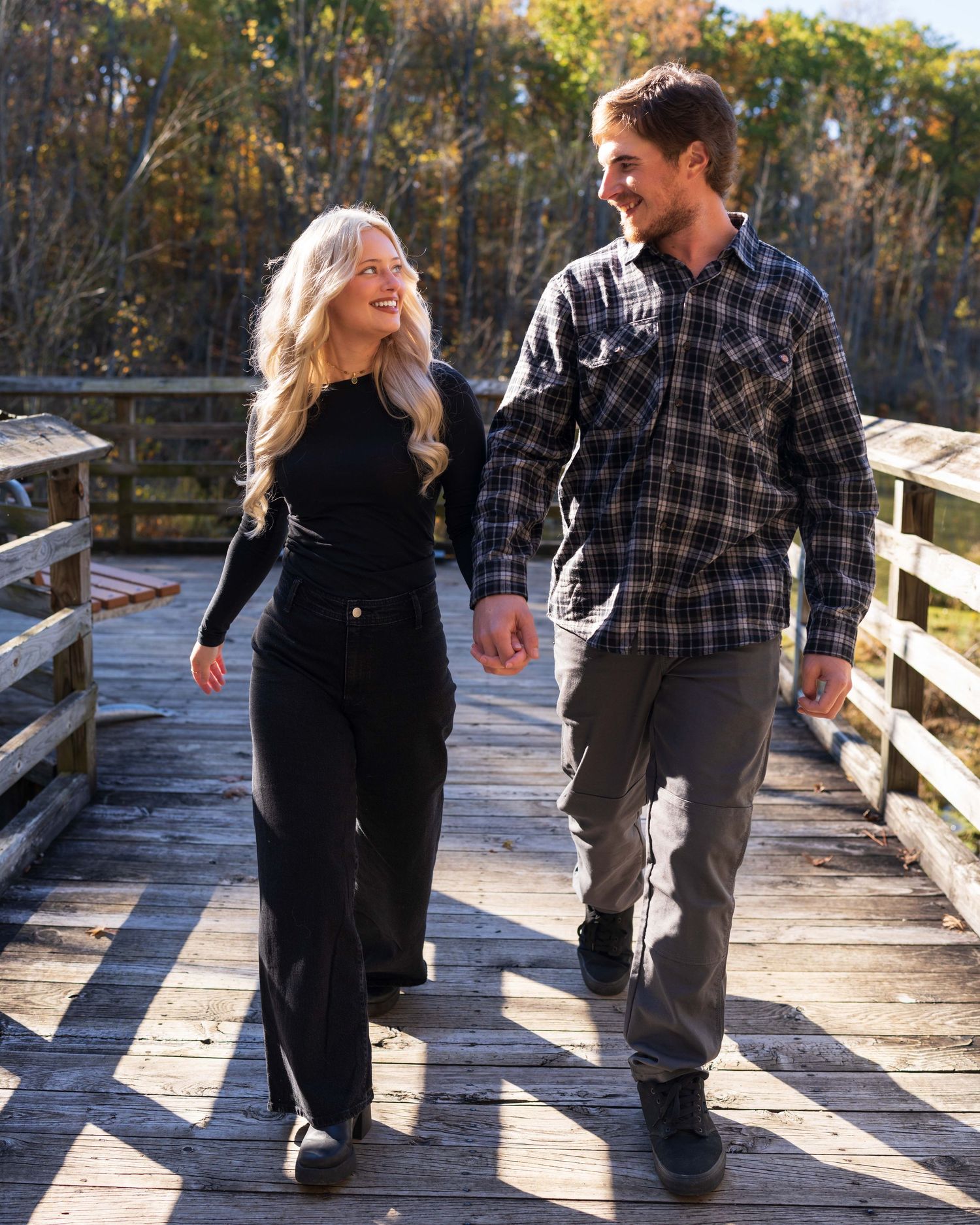 A smiling couple holds hands while walking on a wooden bridge surrounded by colorful autumn foliage.