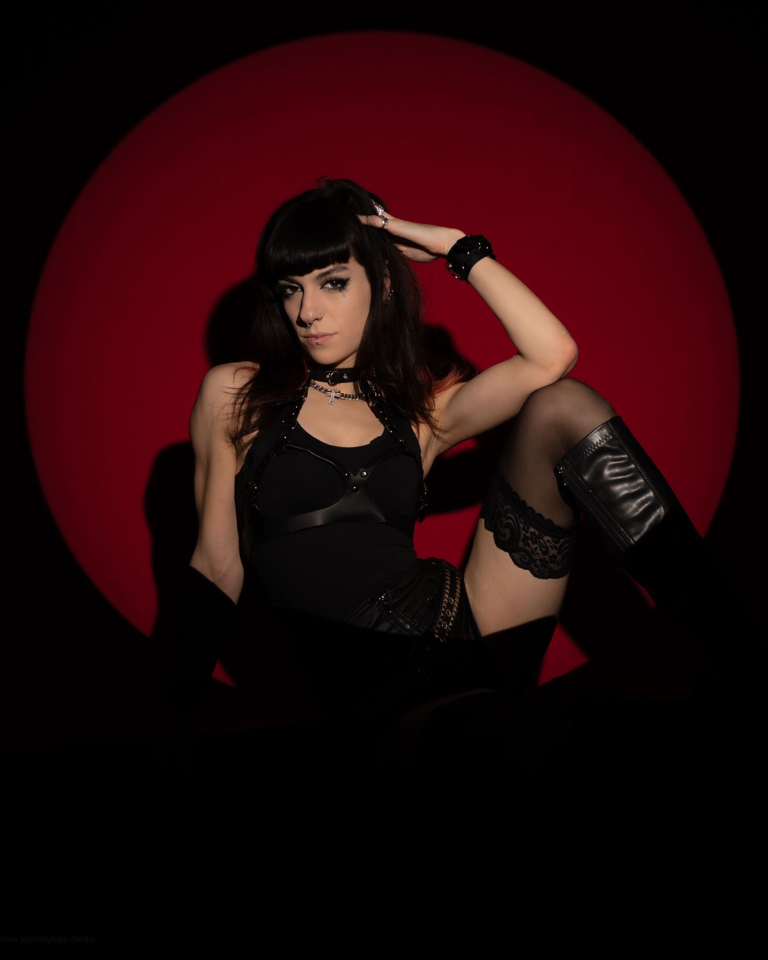 Woman in black lingerie and stockings poses against a dramatic red circle backdrop in a dark, moody setting.