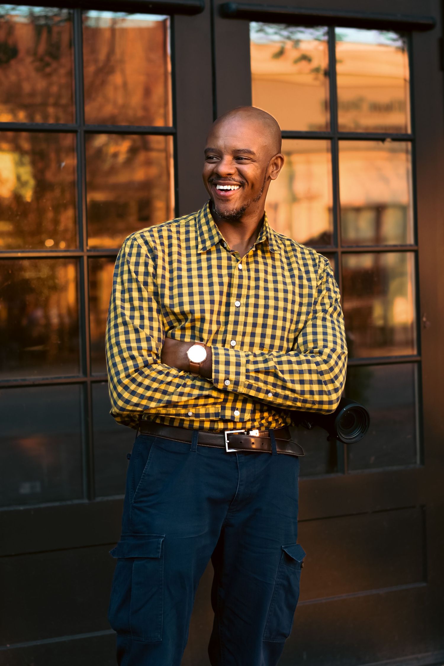 Person in yellow checkered shirt and blue cargo pants poses by window during golden hour lighting.