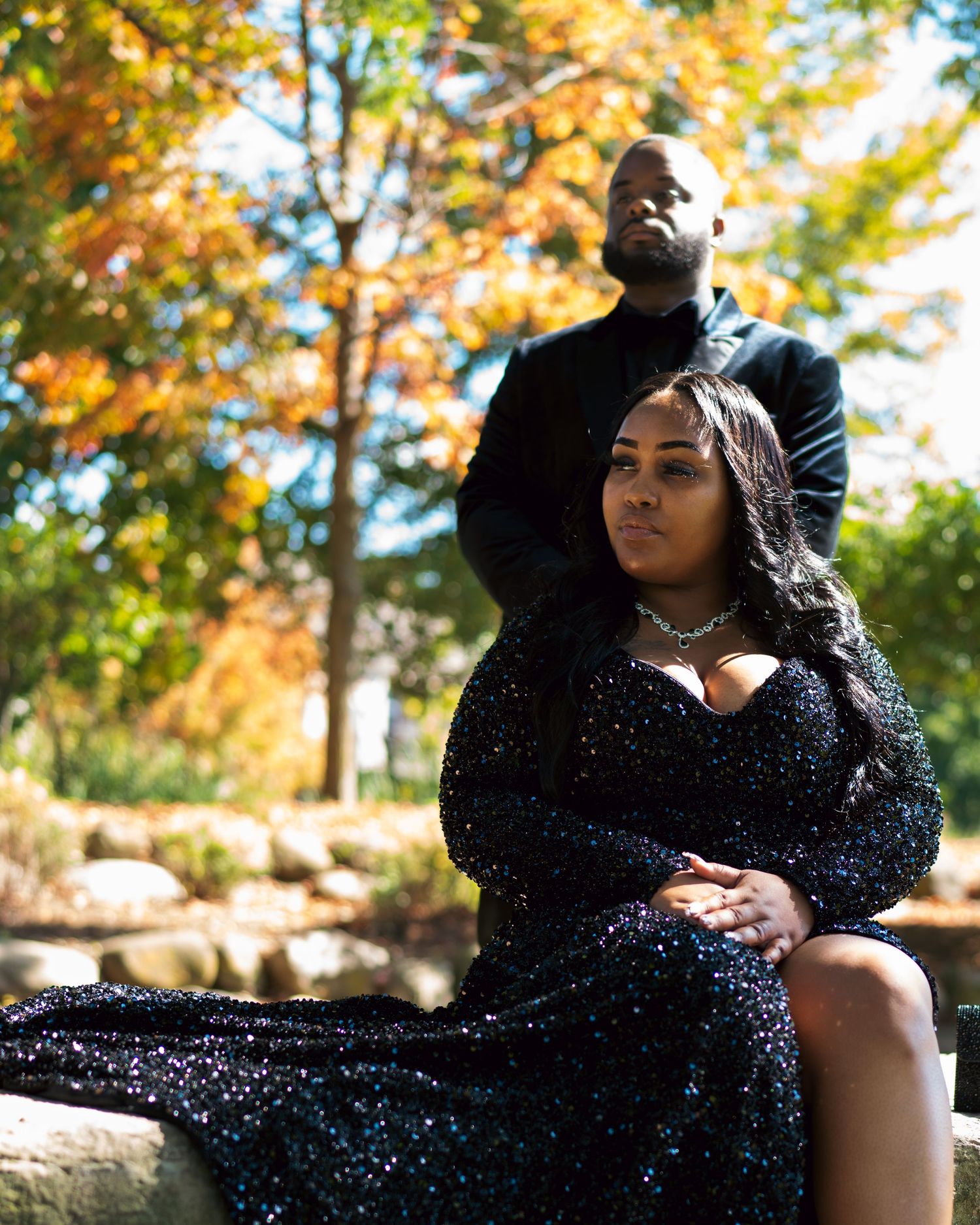 A person in a sparkling black dress poses thoughtfully against an autumn backdrop with golden foliage.