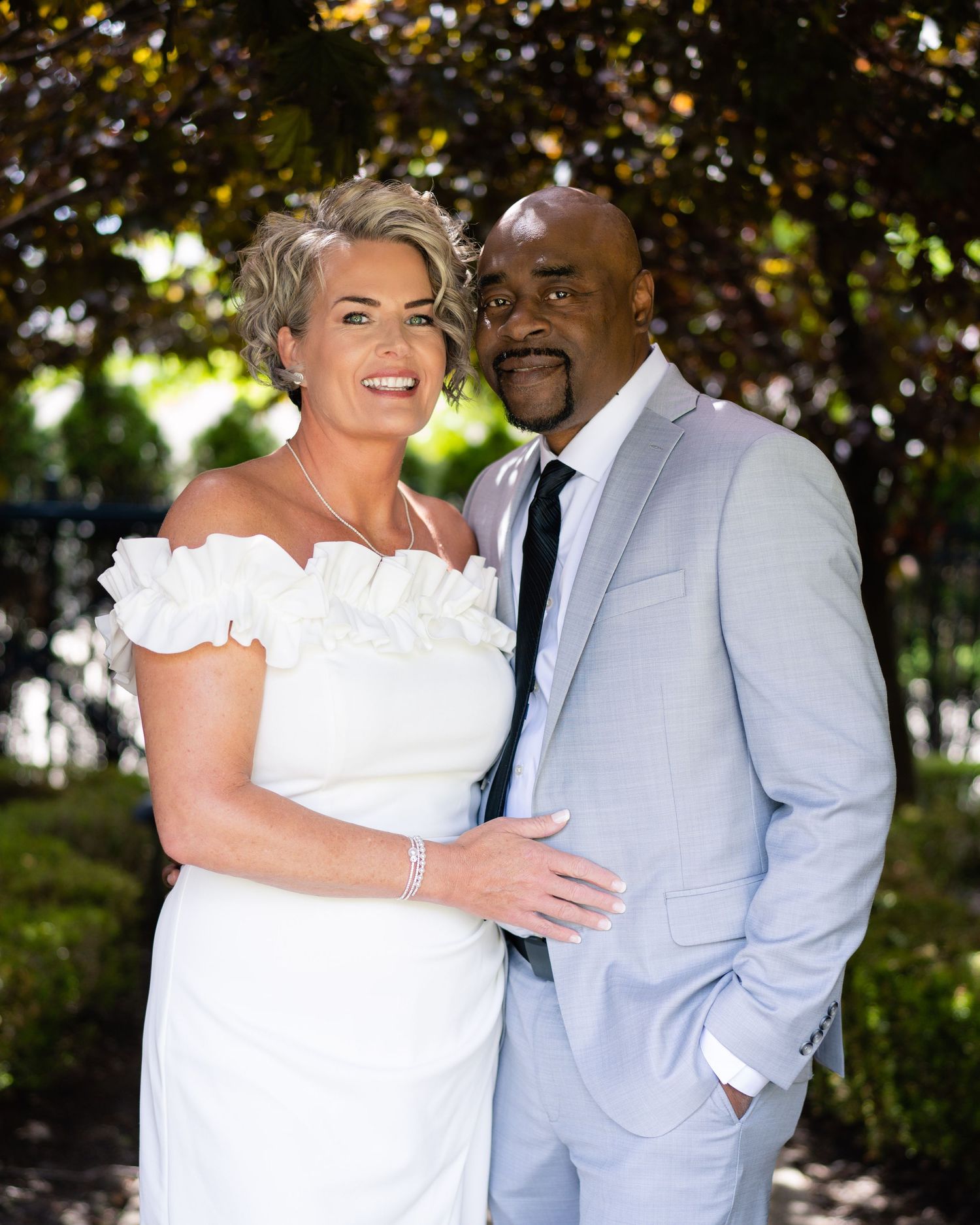Elegant couple posing outdoors, woman in white off-shoulder dress and man in light gray suit, wedding portrait.