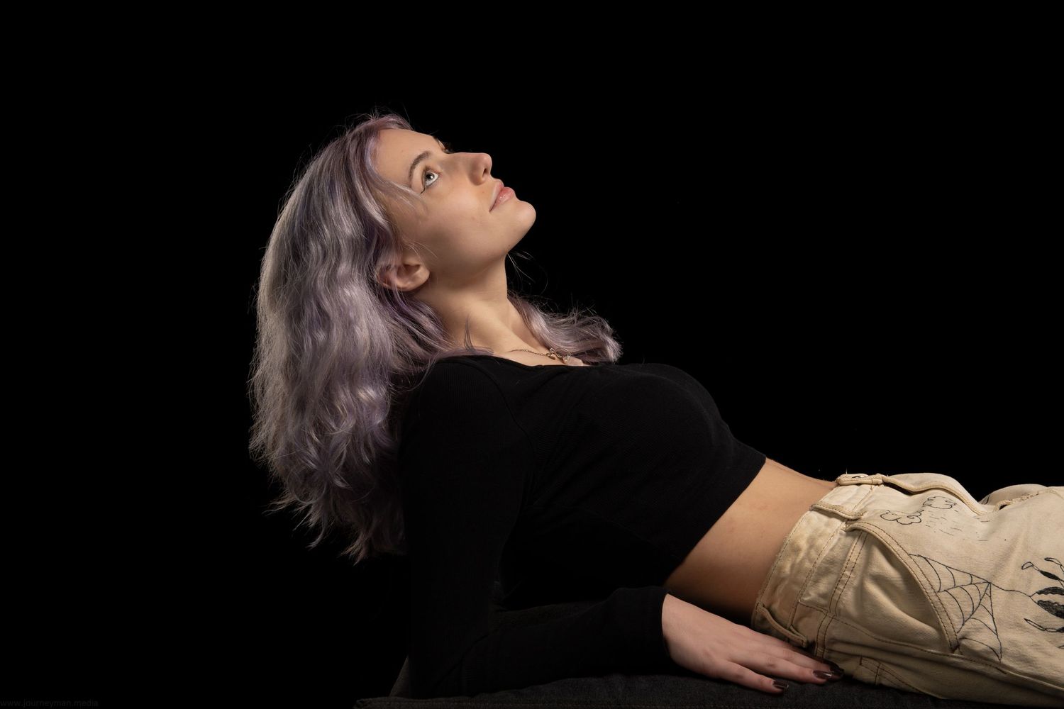 Young woman with silver hair lying down, wearing black top and cargo pants, gazing upward against dark background.
