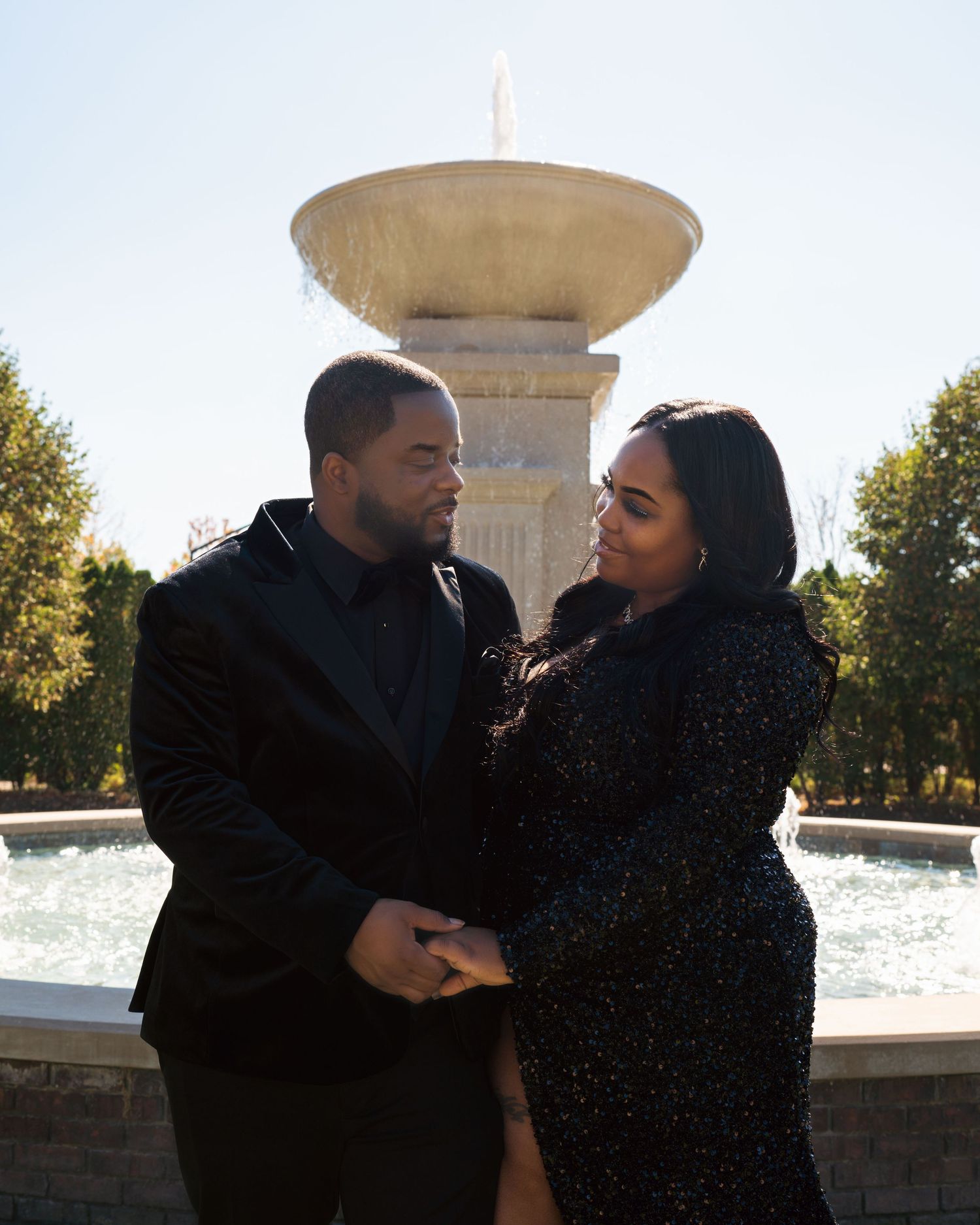 A couple shares romantic moments near a fountain in a park during golden hour lighting.