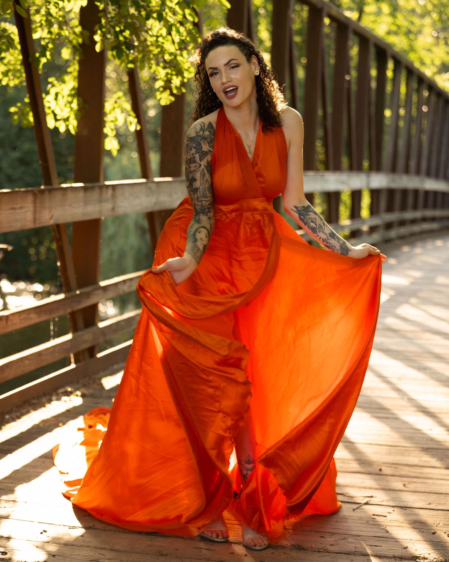 A flowing orange maxi dress twirls on a wooden bridge path amid sunlit trees and greenery.