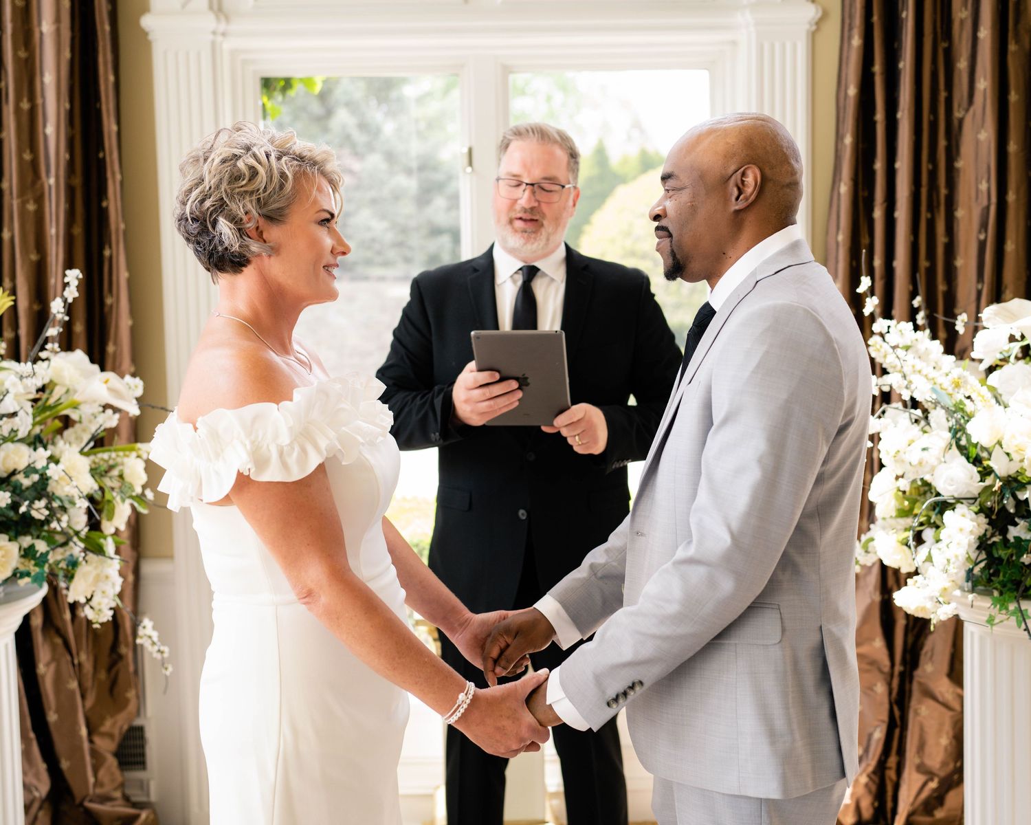 Couple exchanging vows at indoor wedding ceremony with officiant, bride in white gown, groom in gray suit, floral arrangements.