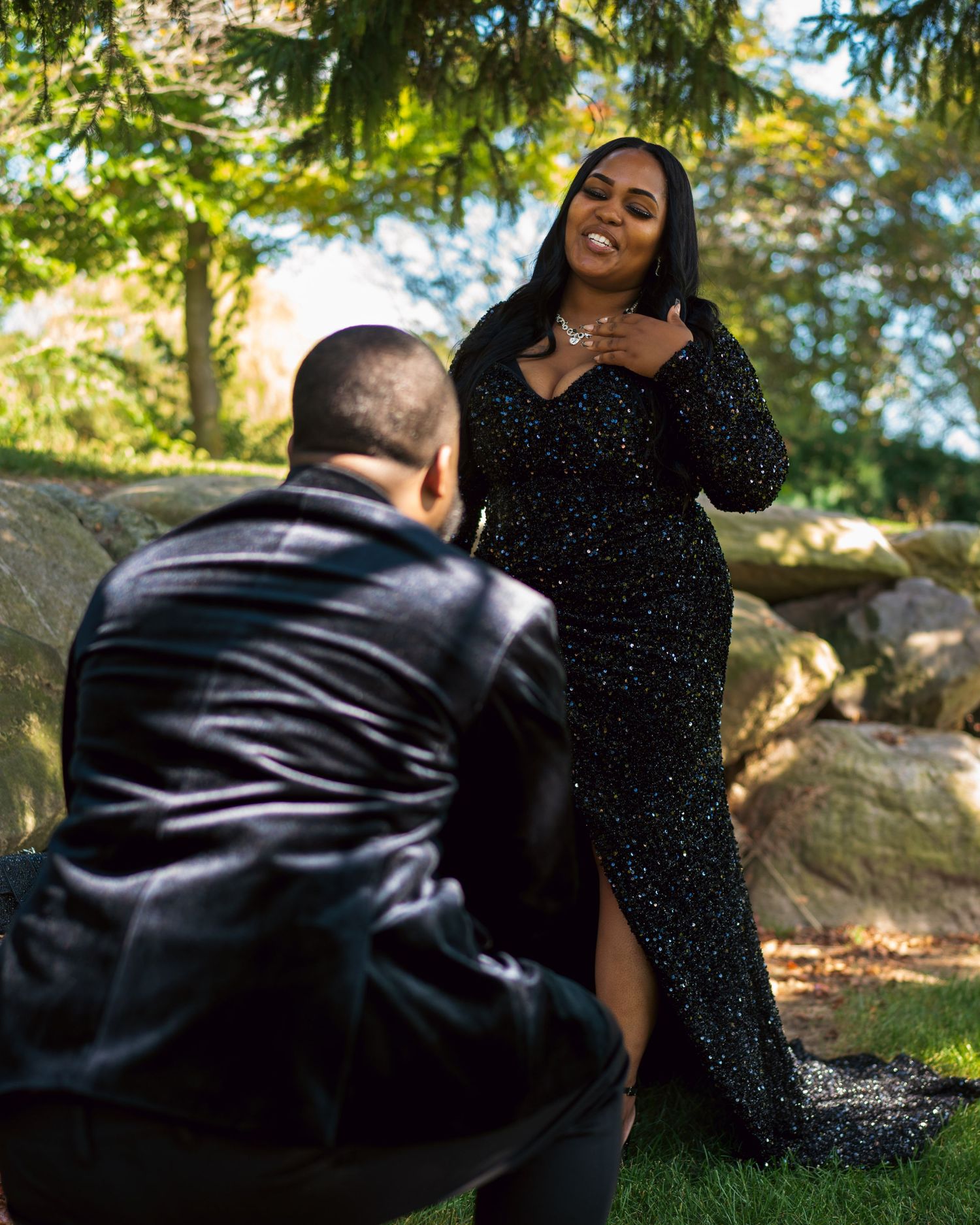 Man kneeling to propose to woman in black sequin gown outdoors near stone wall in lush green garden setting.