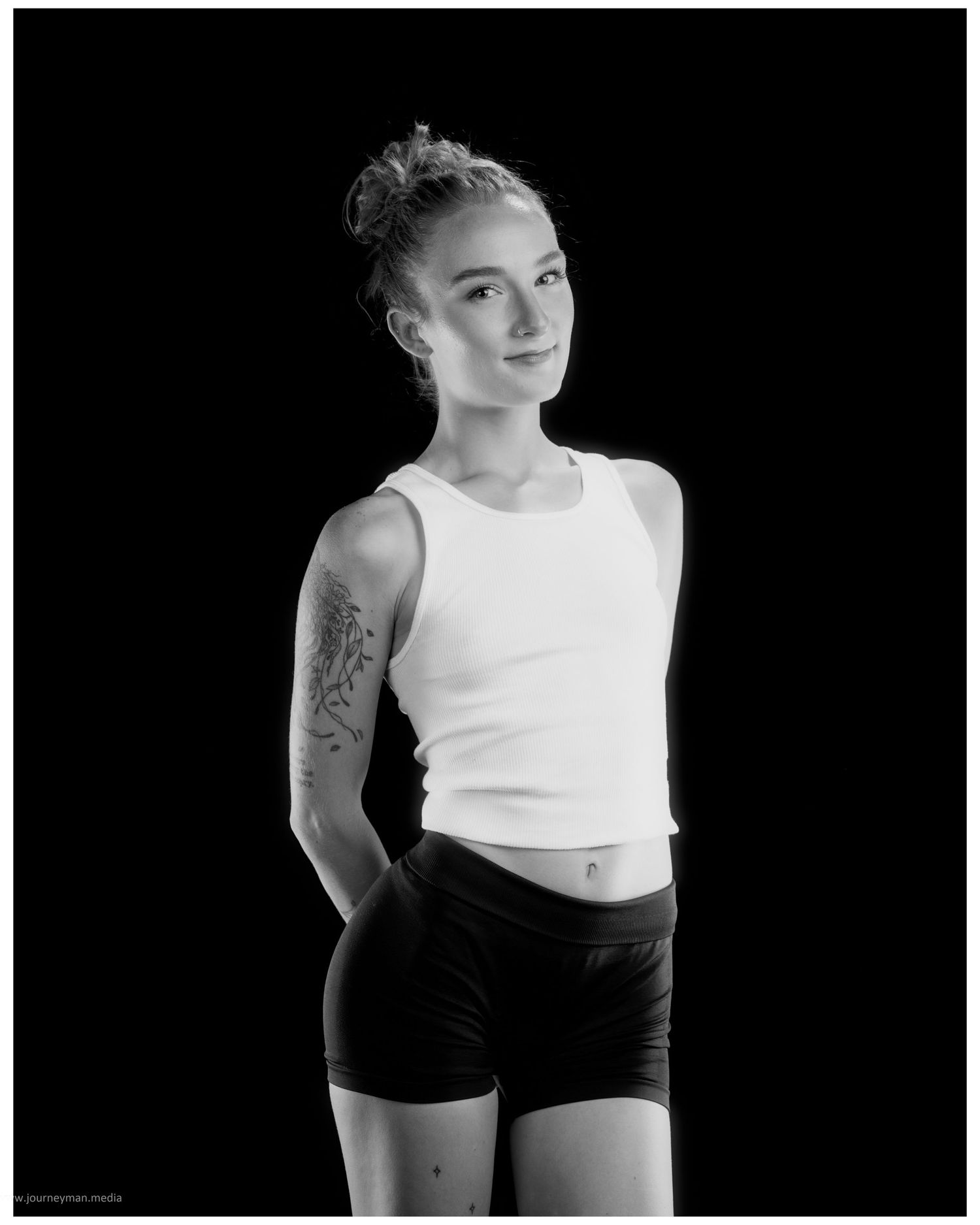 Black and white portrait of a young woman with tattoos wearing a white crop top and black shorts posing against dark background.