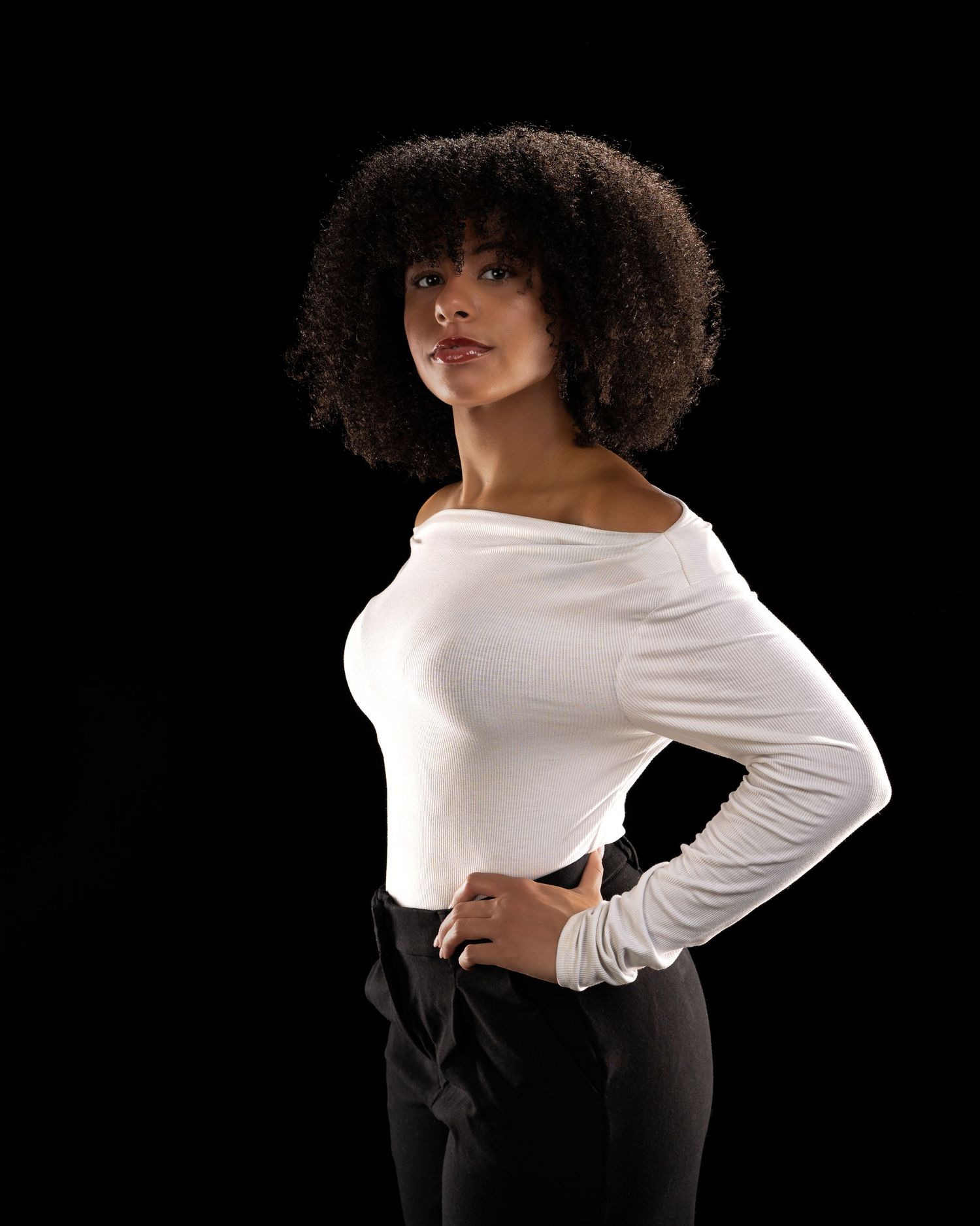 Young woman with curly afro hair poses confidently in white off-shoulder top and black pants against dark background.