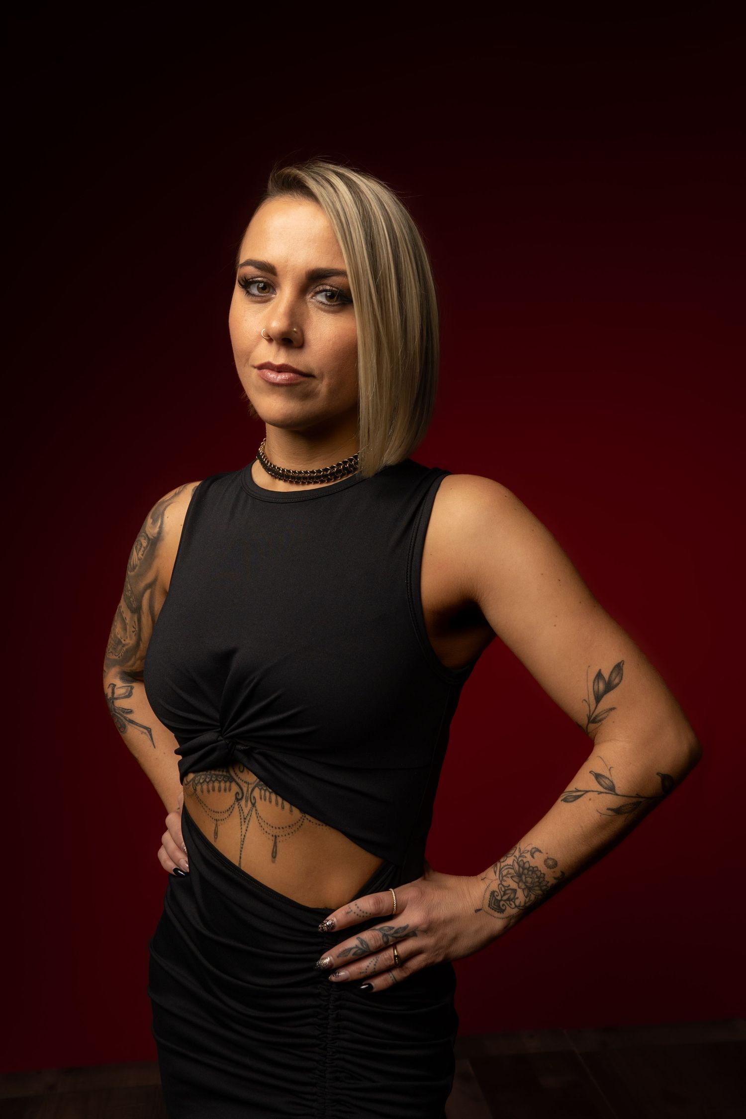 Blonde woman with tattoos wearing black crop top and skirt poses confidently against dark red studio background.