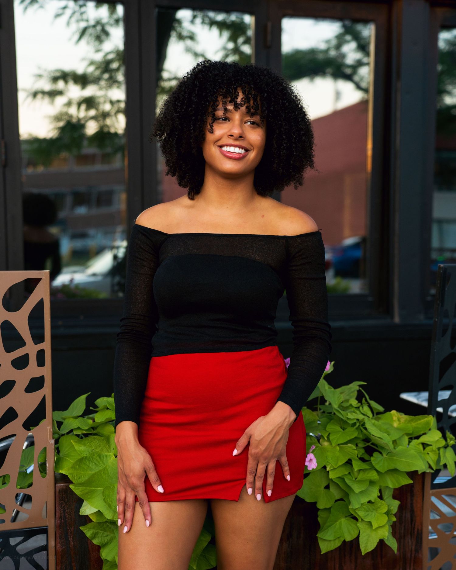 A woman in a black off-shoulder top and red shorts poses outdoors near green plants and decorative wall elements.