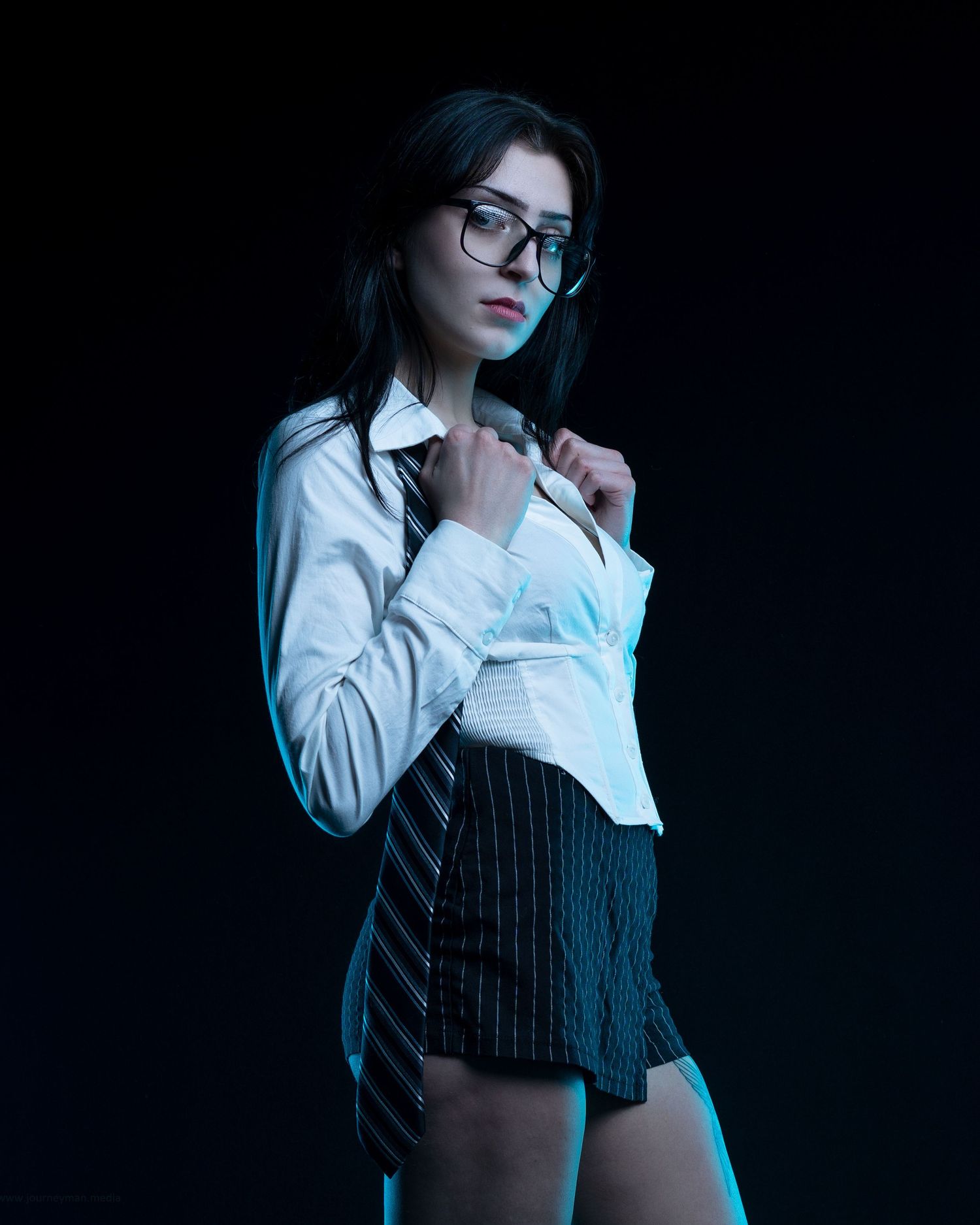 Woman in white shirt, tie, mini skirt and glasses poses against dark background in stylish editorial look.