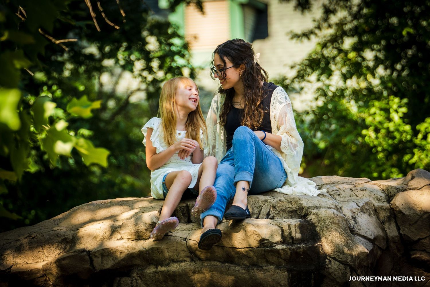 A woman and young girl sit on rocks outdoors, smiling and talking in a sunlit garden setting.