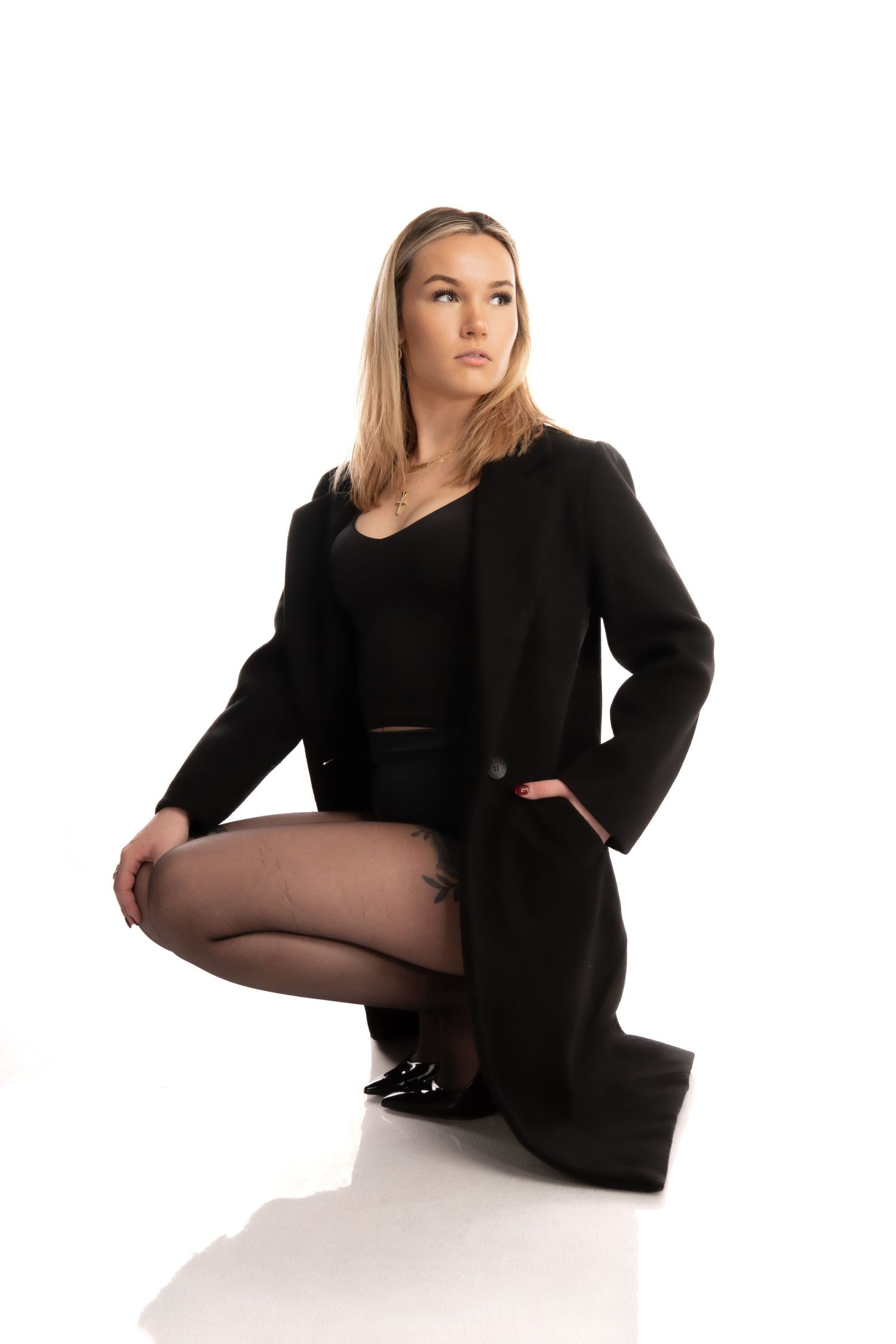 Fashion model in black coat and bodysuit poses crouching on white background in studio setting.
