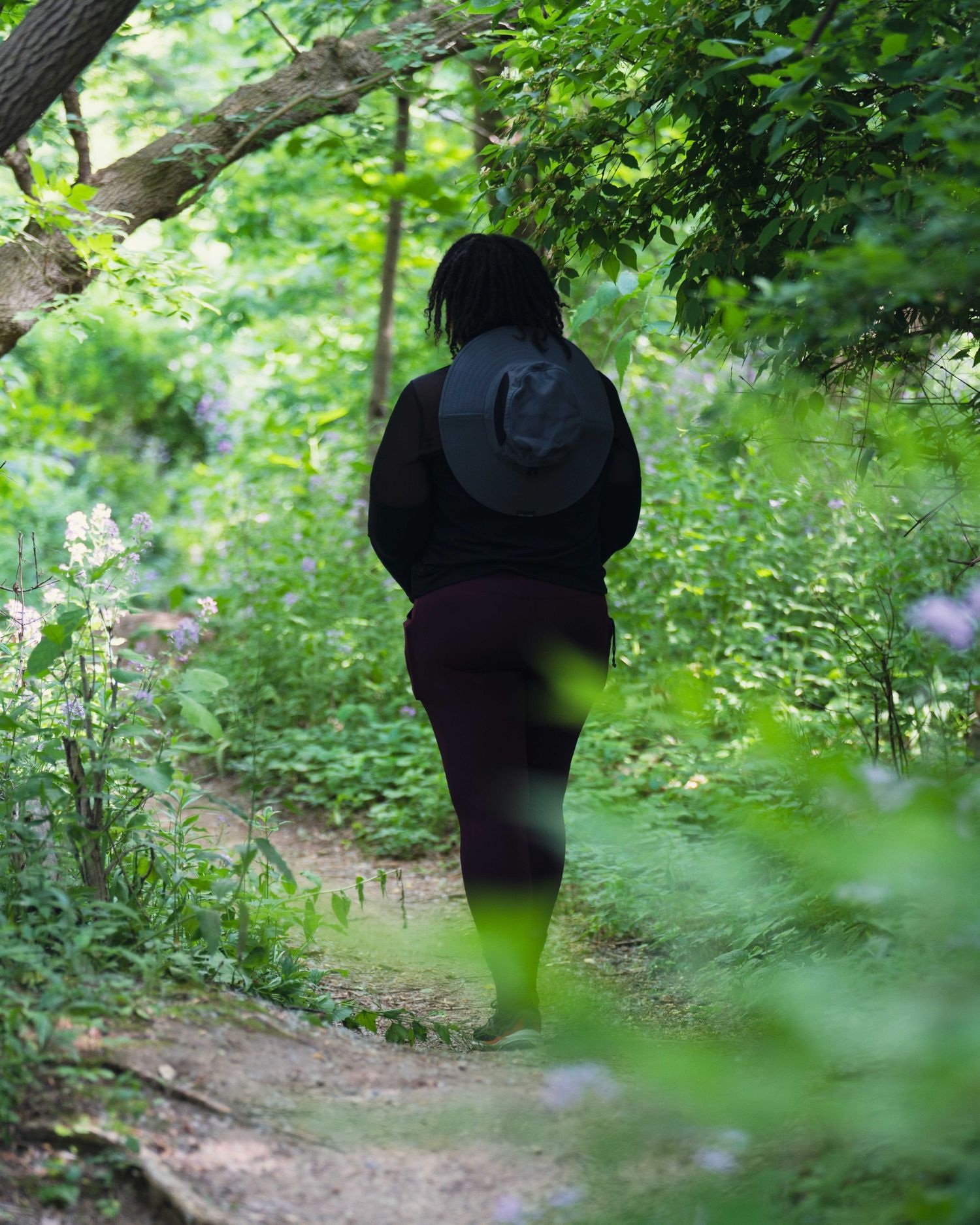 A person in dark clothing walks along a winding forest trail surrounded by lush green vegetation and dappled sunlight.