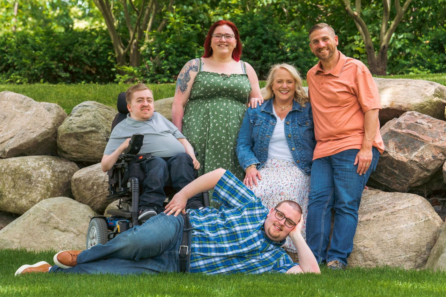 Family of five posing outdoors near large rocks, including a person in a wheelchair, smiling together on green grass.