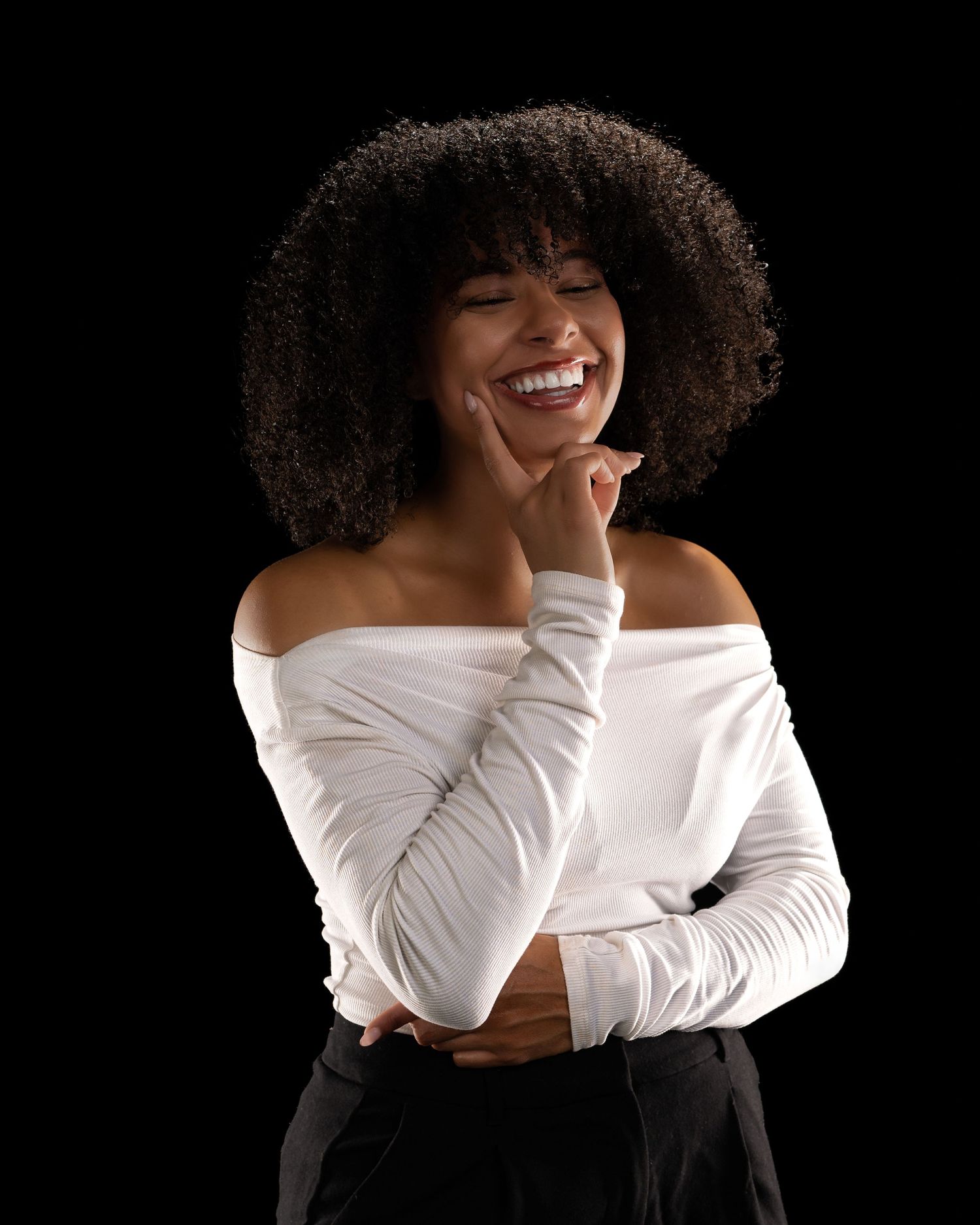 A young Black woman with curly hair laughs joyfully, wearing a white off-shoulder top against a black background.