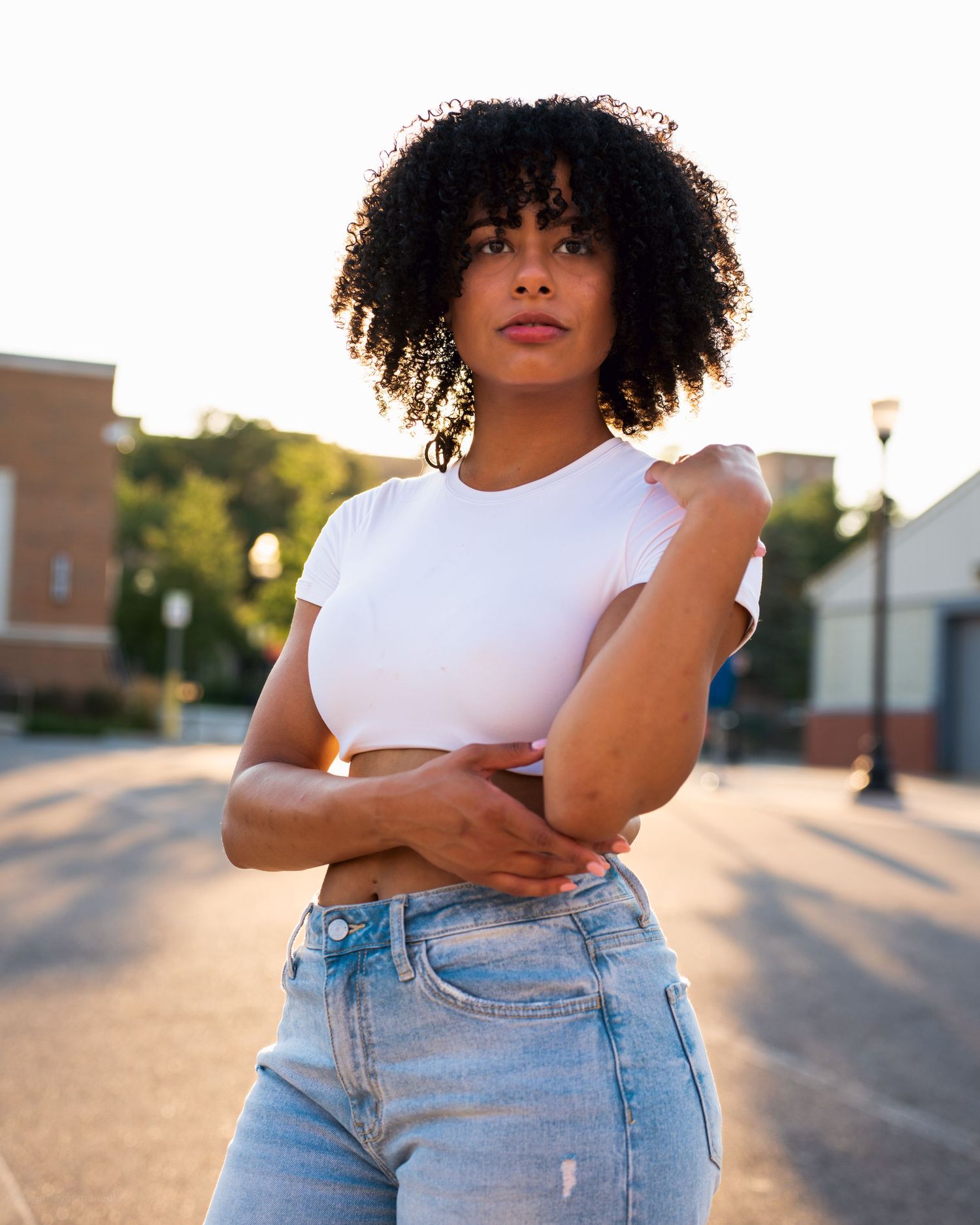 A woman in a white crop top and high waist jeans poses outdoors at sunset.