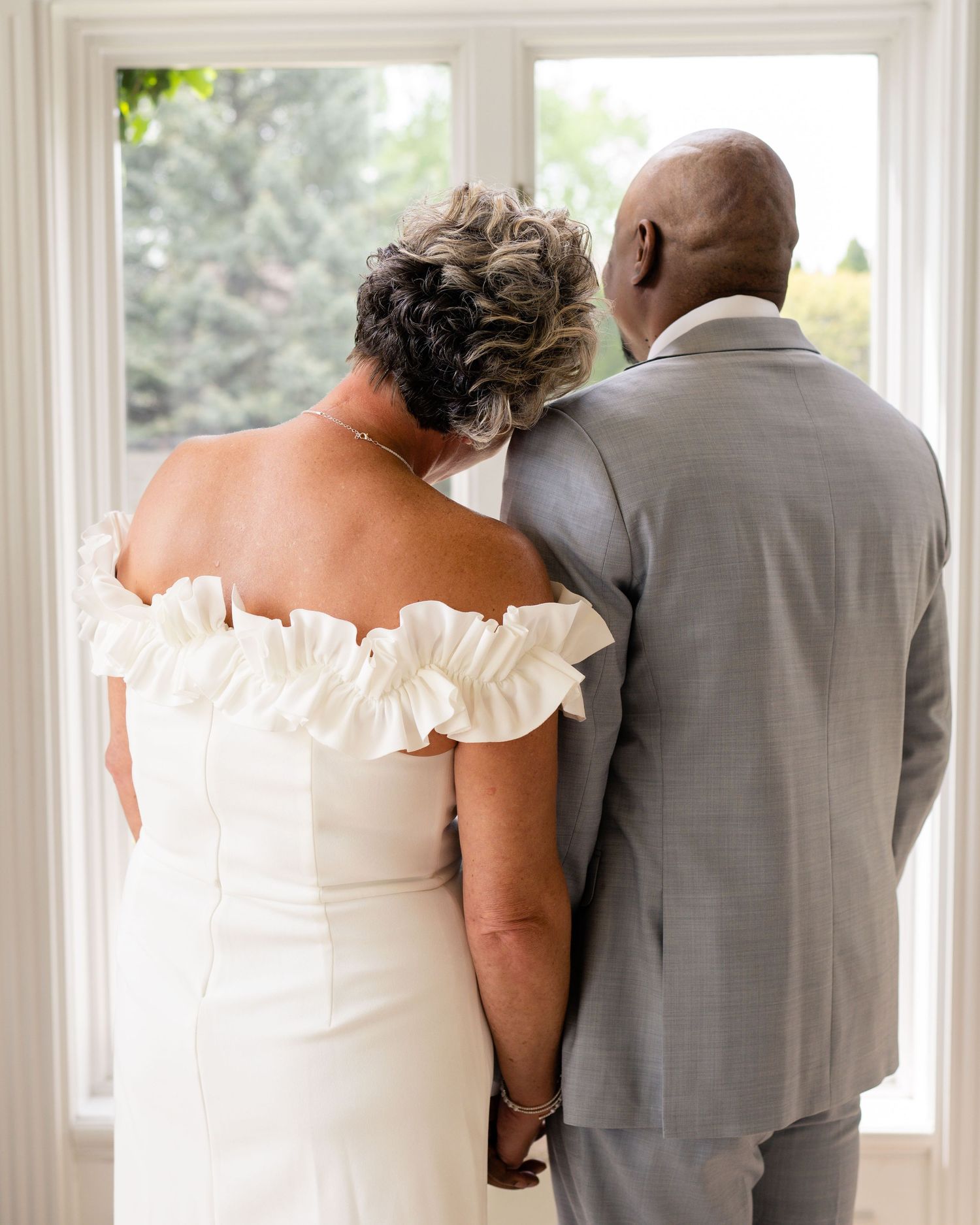 Bride in off-shoulder white gown and groom in gray suit hold hands, looking out a window together.