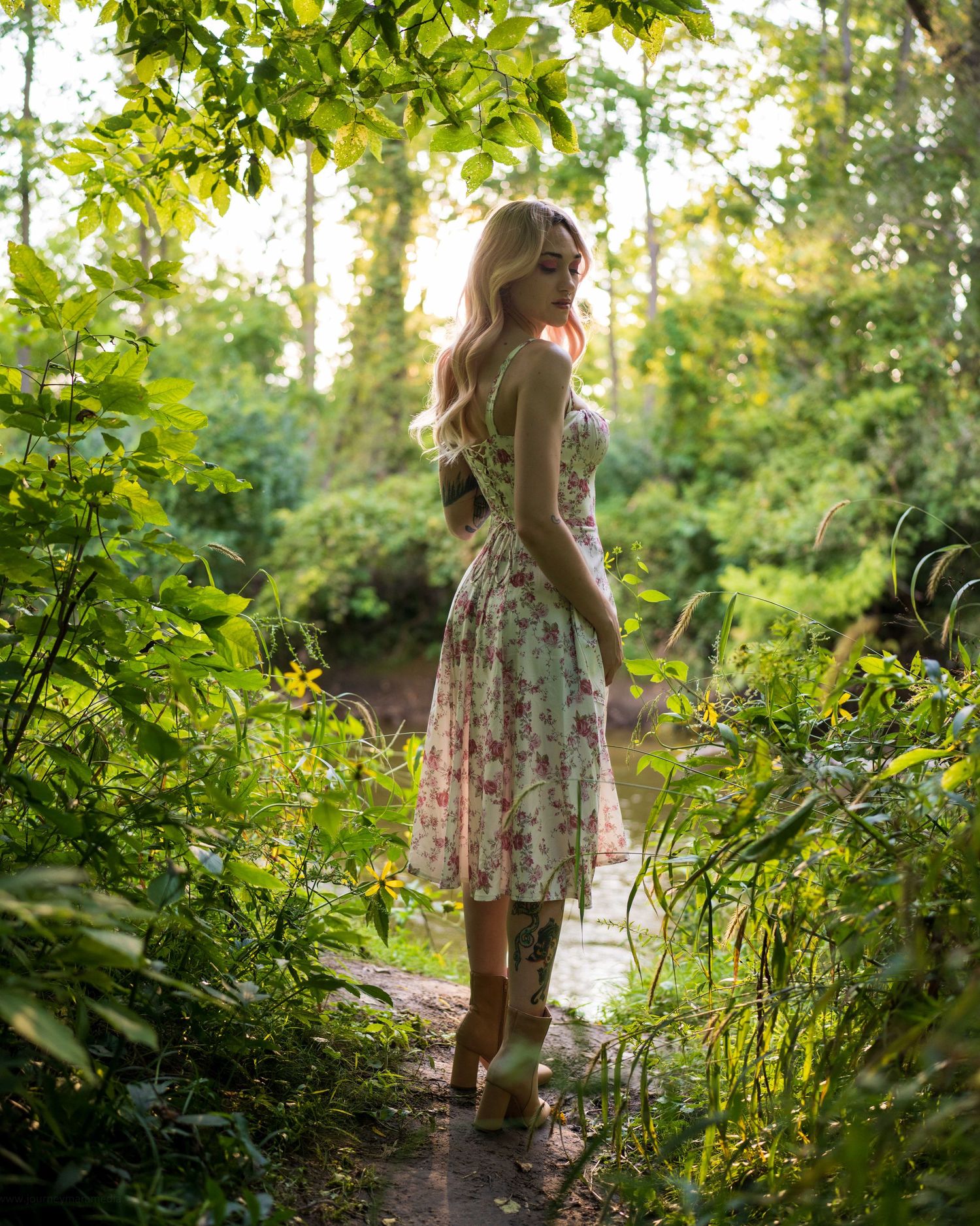 Woman in floral dress standing on garden path surrounded by lush green trees in golden sunlight.
