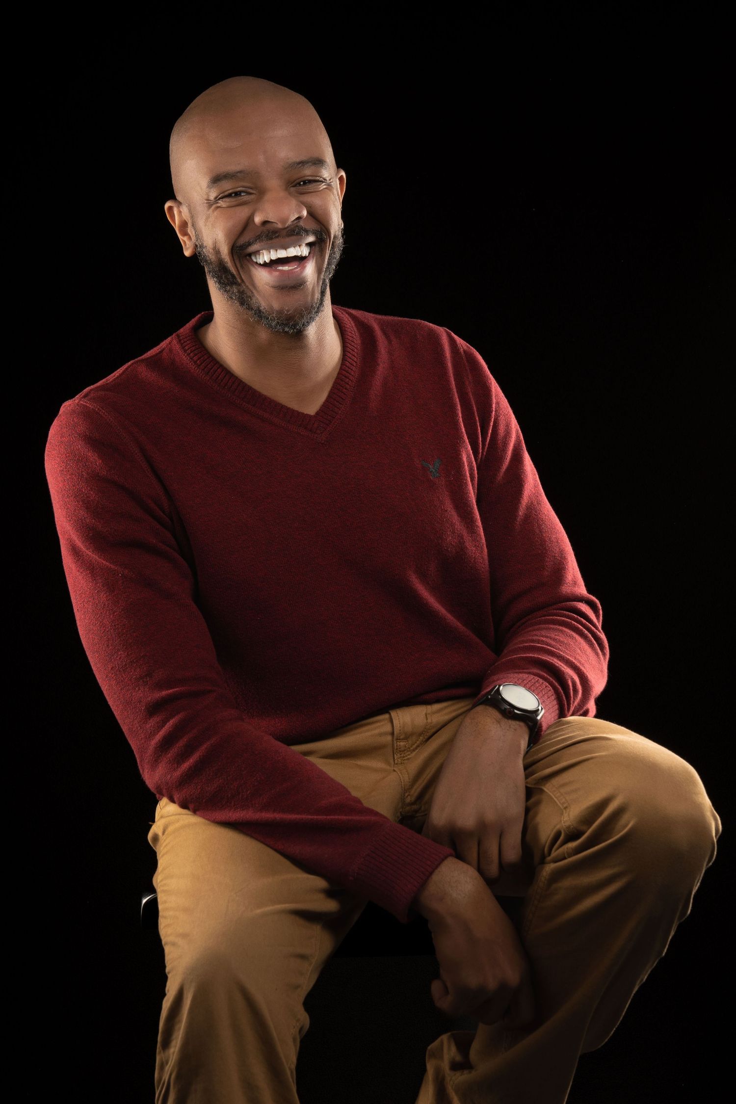 Man in burgundy v-neck sweater and khaki pants smiles warmly while seated against black background.