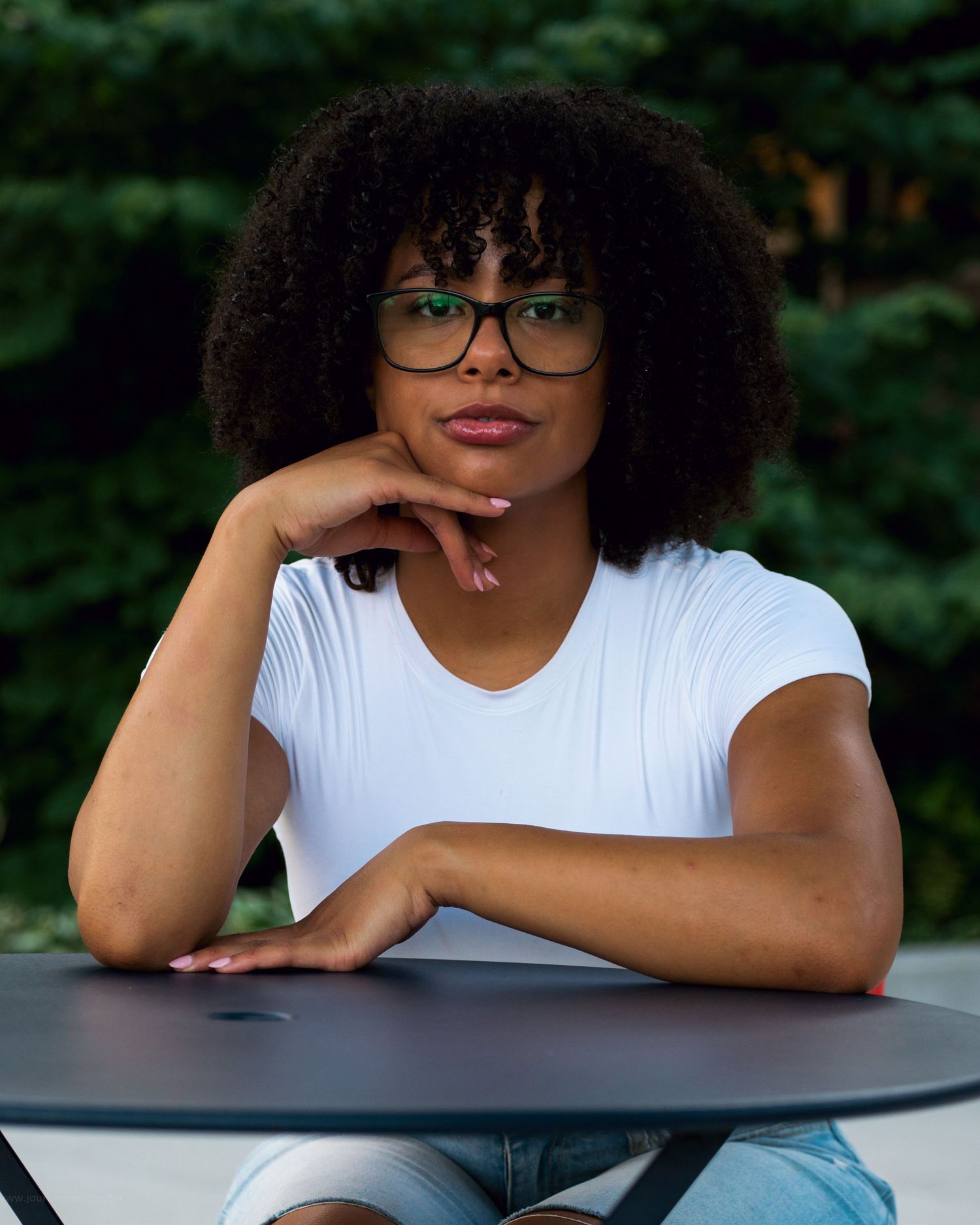 A person wearing glasses in a white t-shirt and curly hair leans on an outdoor table in a thoughtful pose.