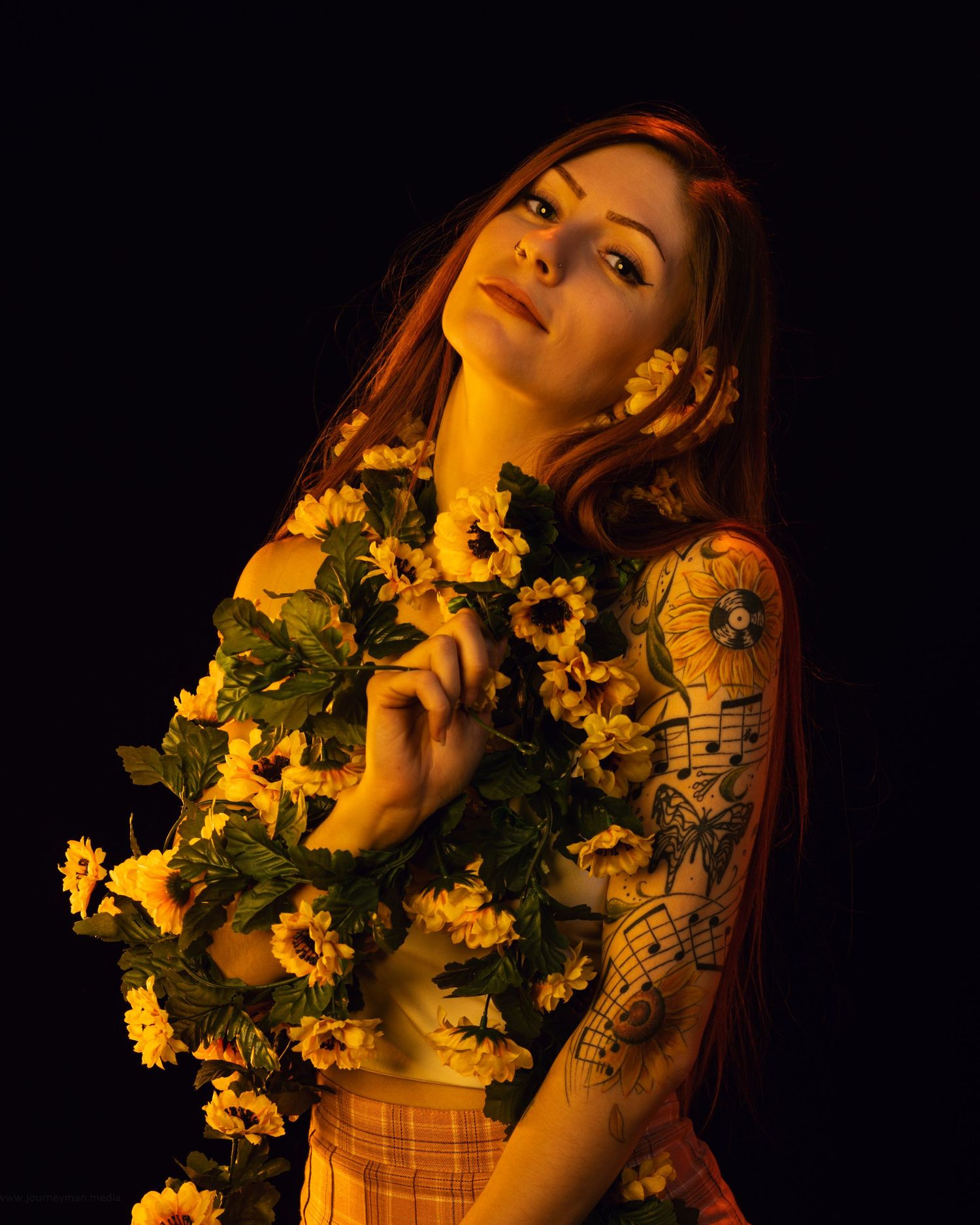 Woman with red hair and tattoos holding yellow sunflowers against a dark background in dramatic warm lighting.