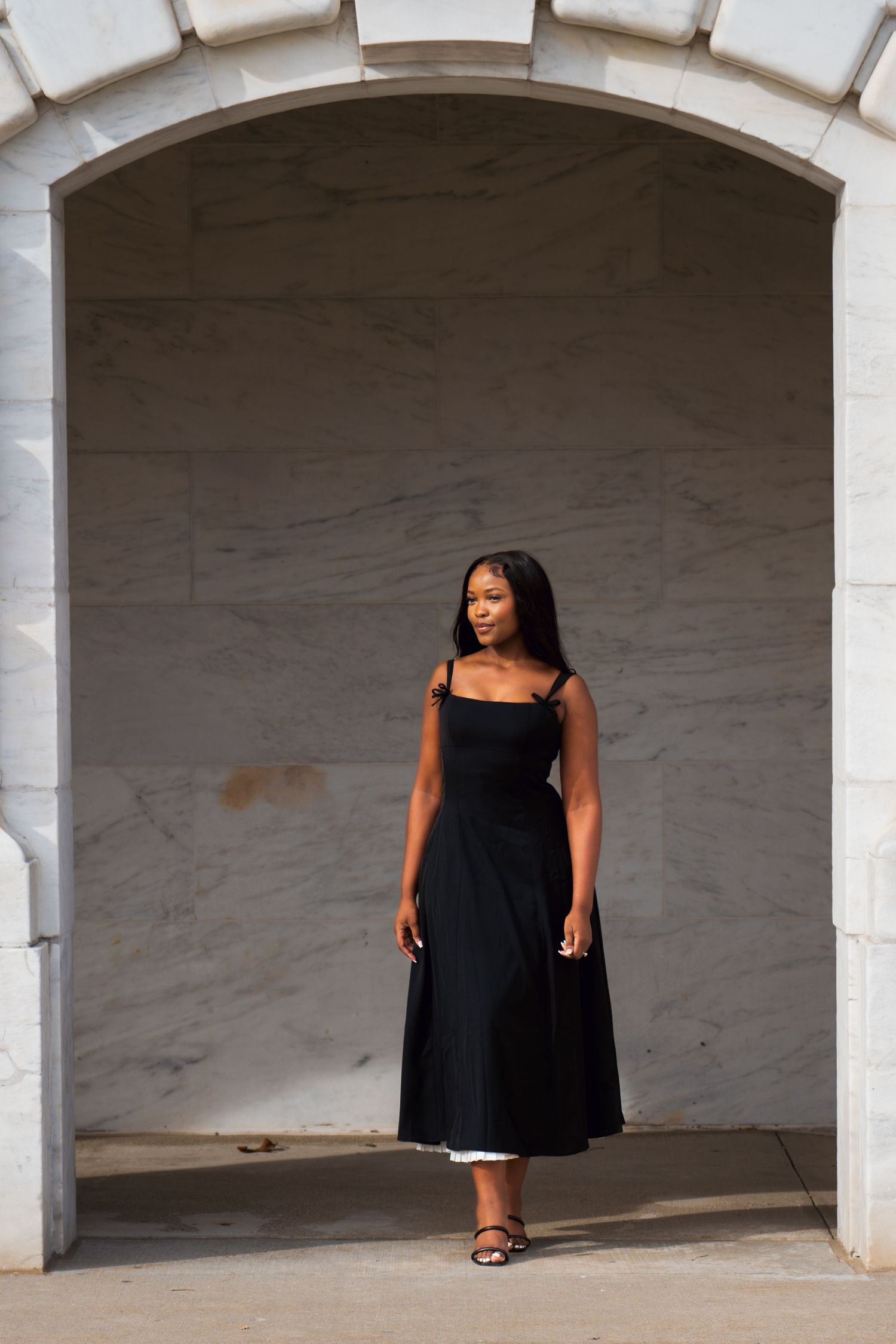 Plus size fashion model wearing black sundress poses in series of architectural archways against white concrete wall.