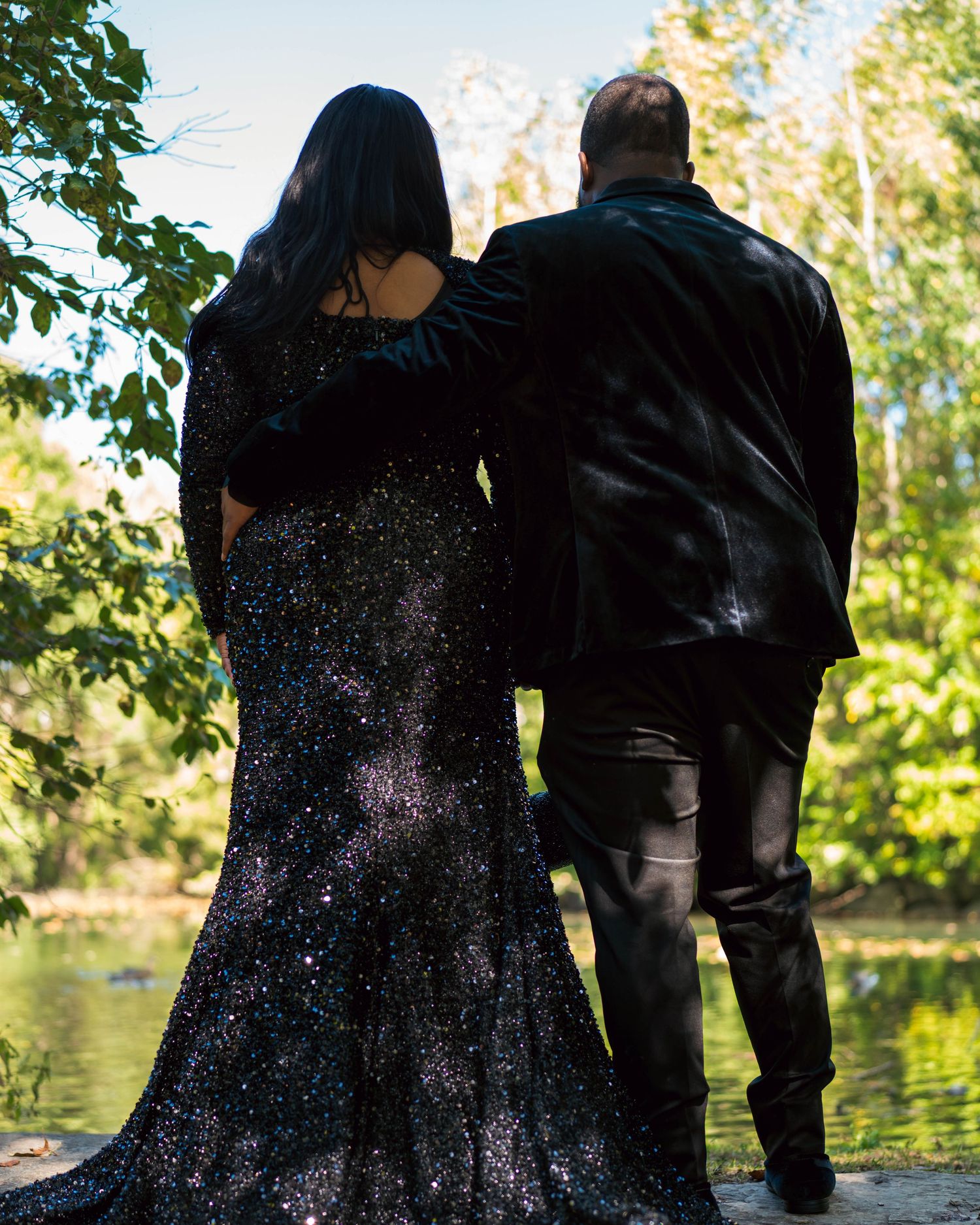 A couple in formal attire poses together by a lake with trees and natural scenery in the background.