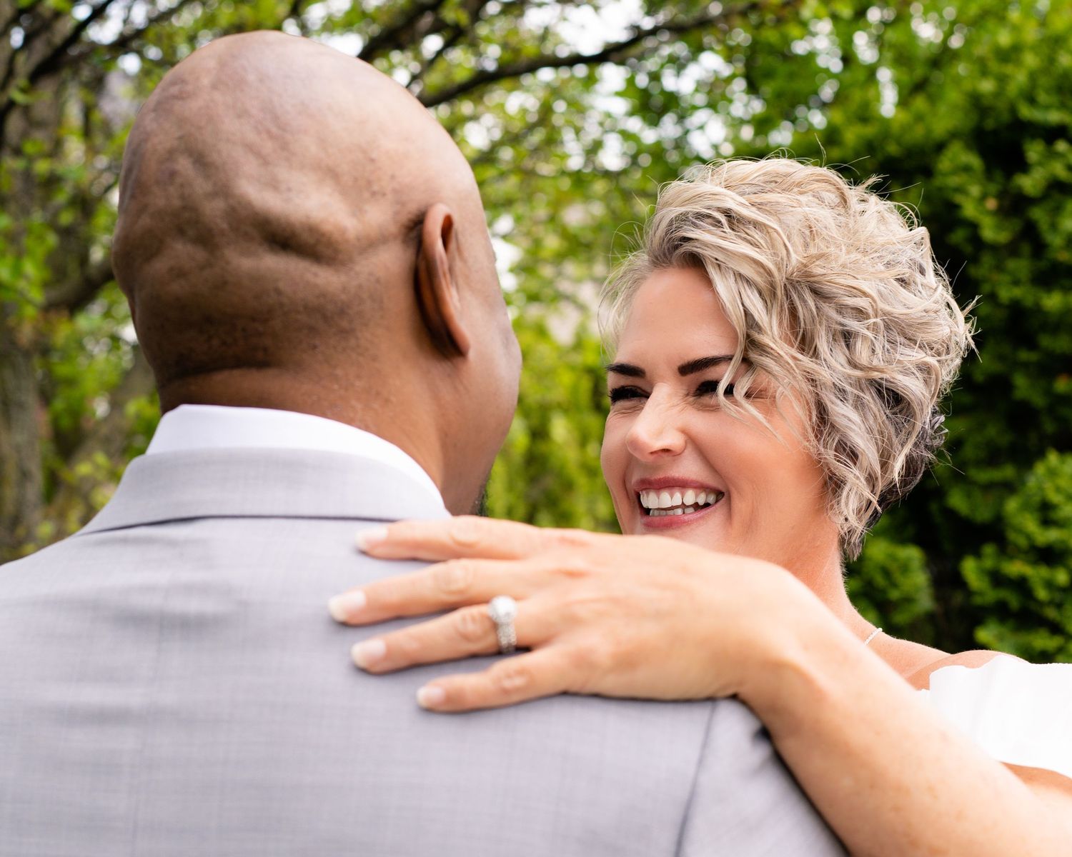 Smiling bride with short blonde hair embraces groom in gray suit outdoors, showing diamond engagement ring.