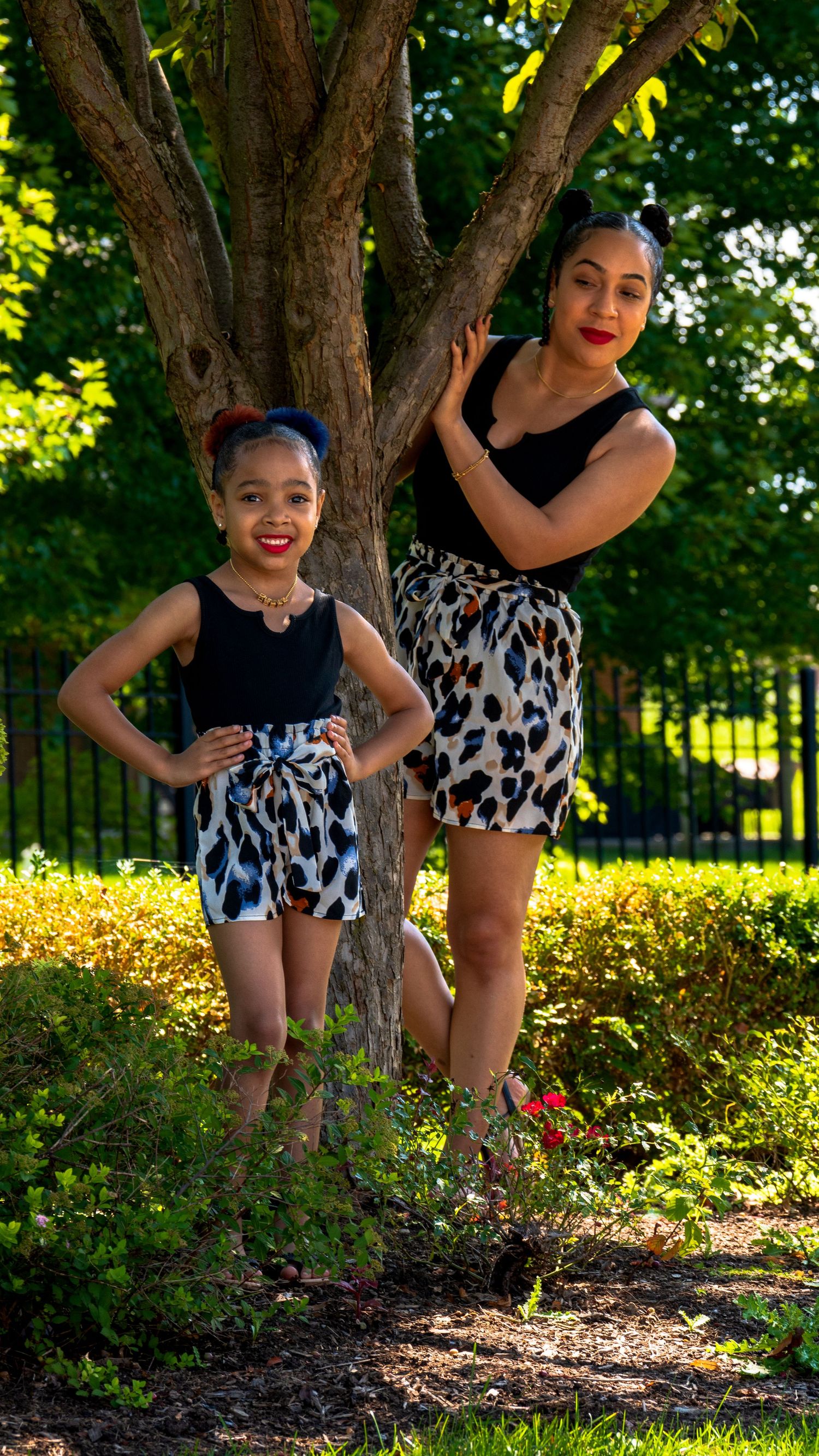 A woman and young girl in matching black tops and leopard print shorts pose together by a tree in a lush green park.