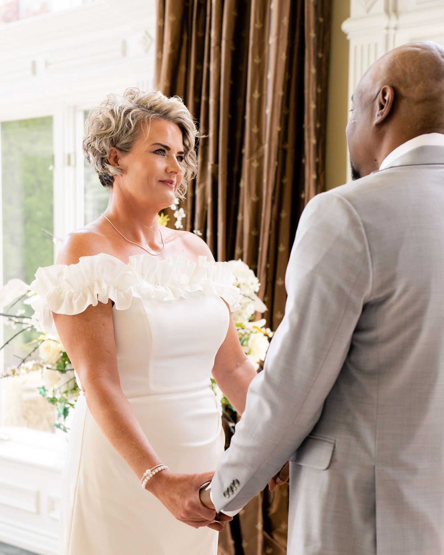 Bride in white off-shoulder gown holds hands with groom in gray suit during intimate wedding ceremony.