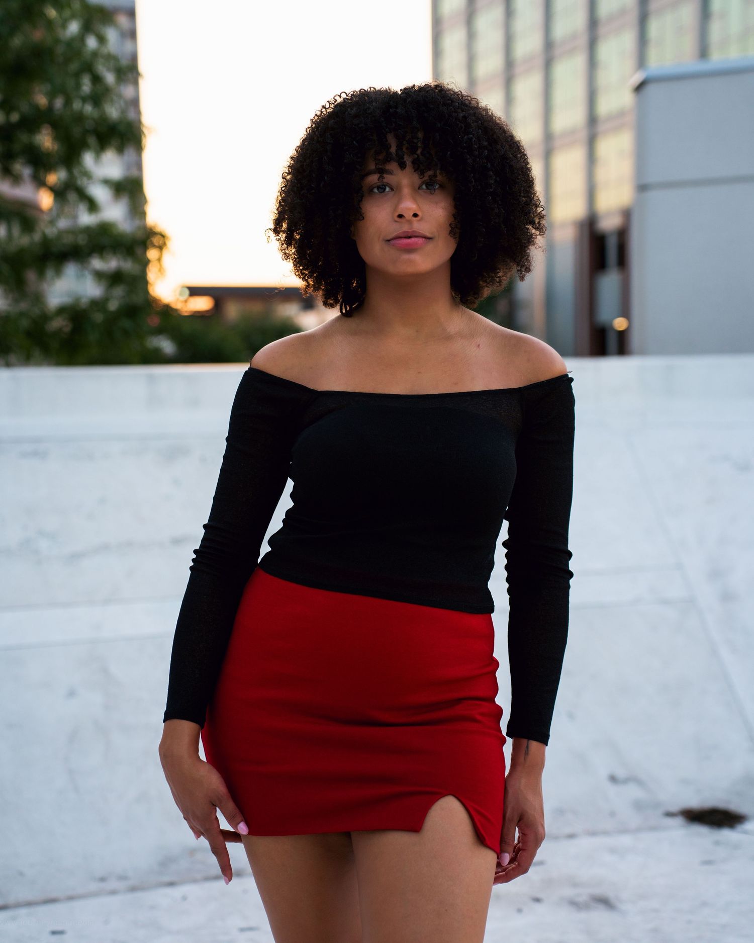 A woman in a black long-sleeve top and red mini skirt poses outdoors against a white wall at sunset.