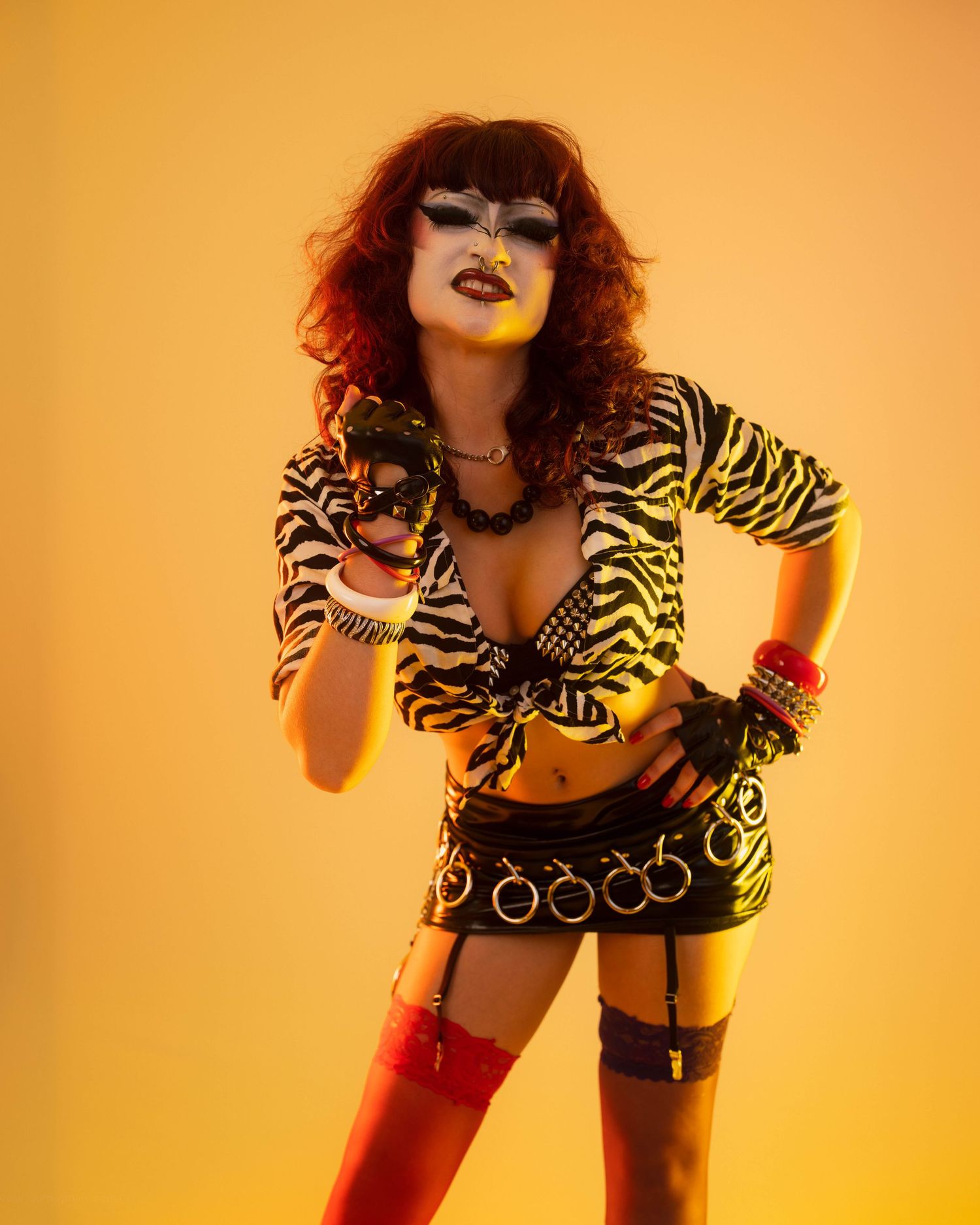 Fierce performer in zebra print top, black mini skirt, red boots, and mask poses on yellow background.