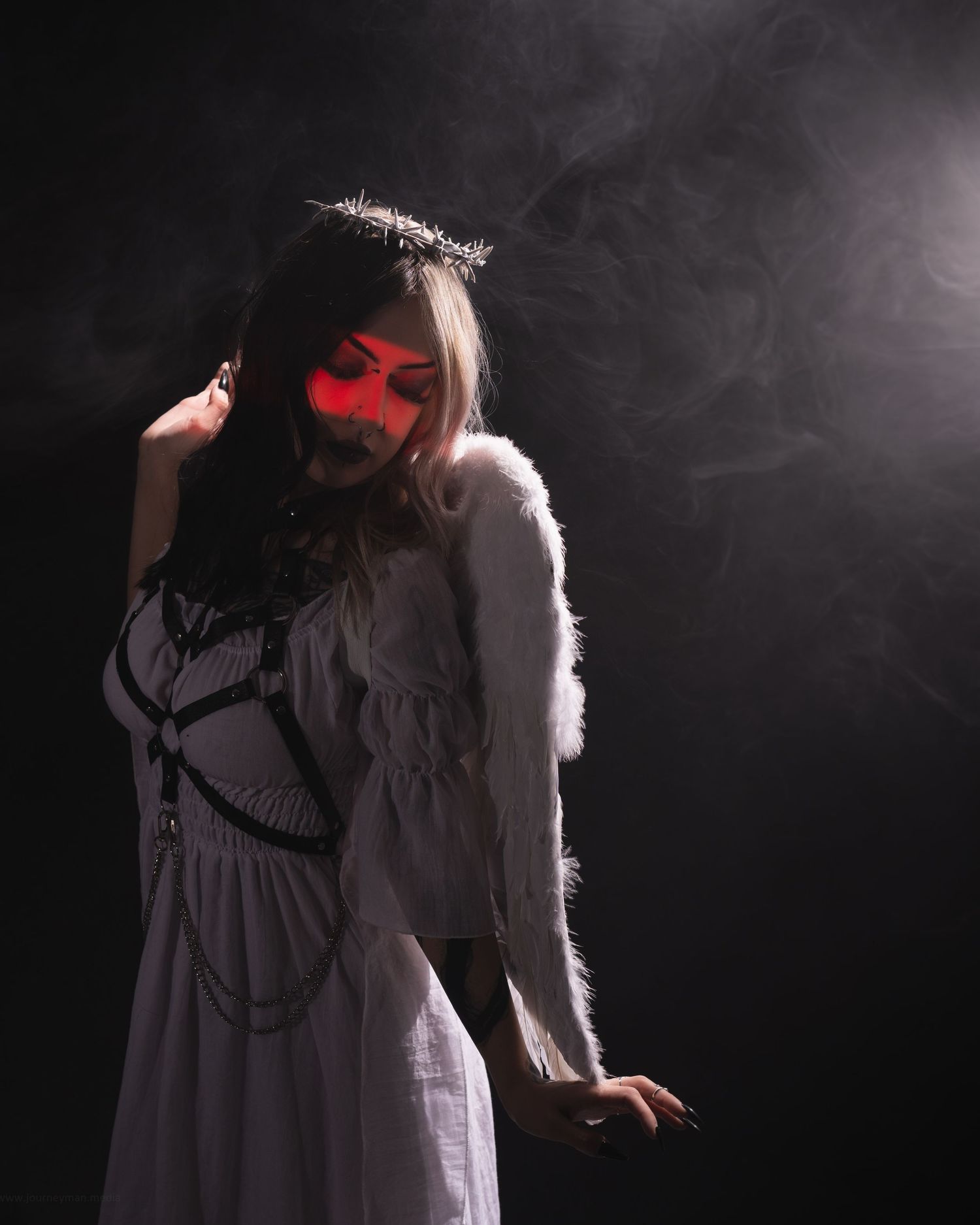 Dark angel cosplay with white dress, feather wings, crown, and red face paint against smoky black background.
