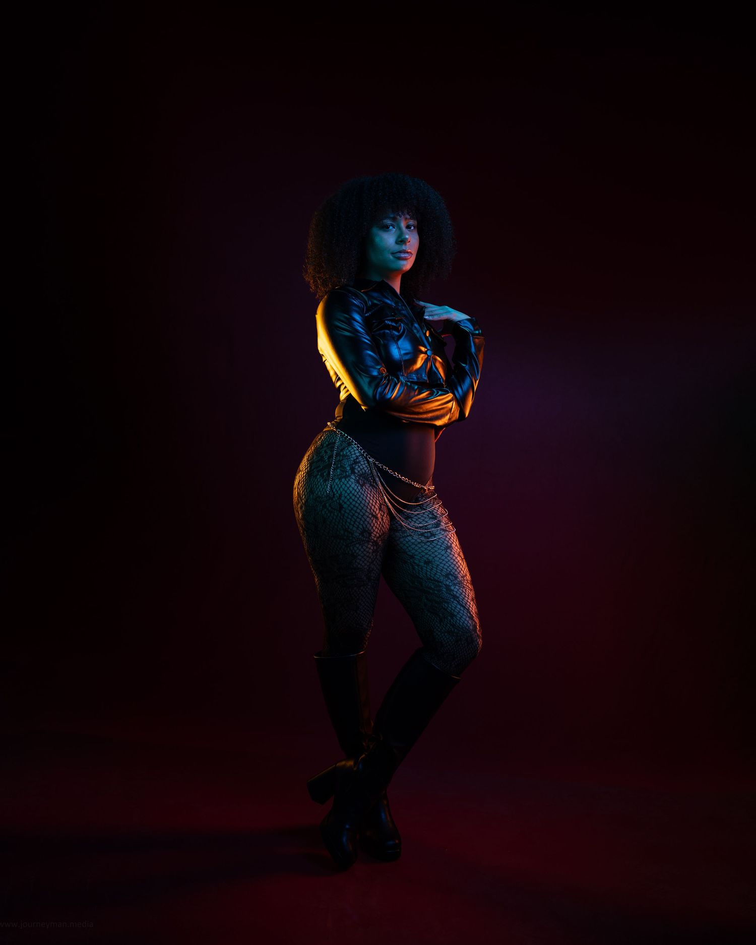 A woman in a leather jacket and jeans poses confidently under dramatic neon lighting against a dark background.
