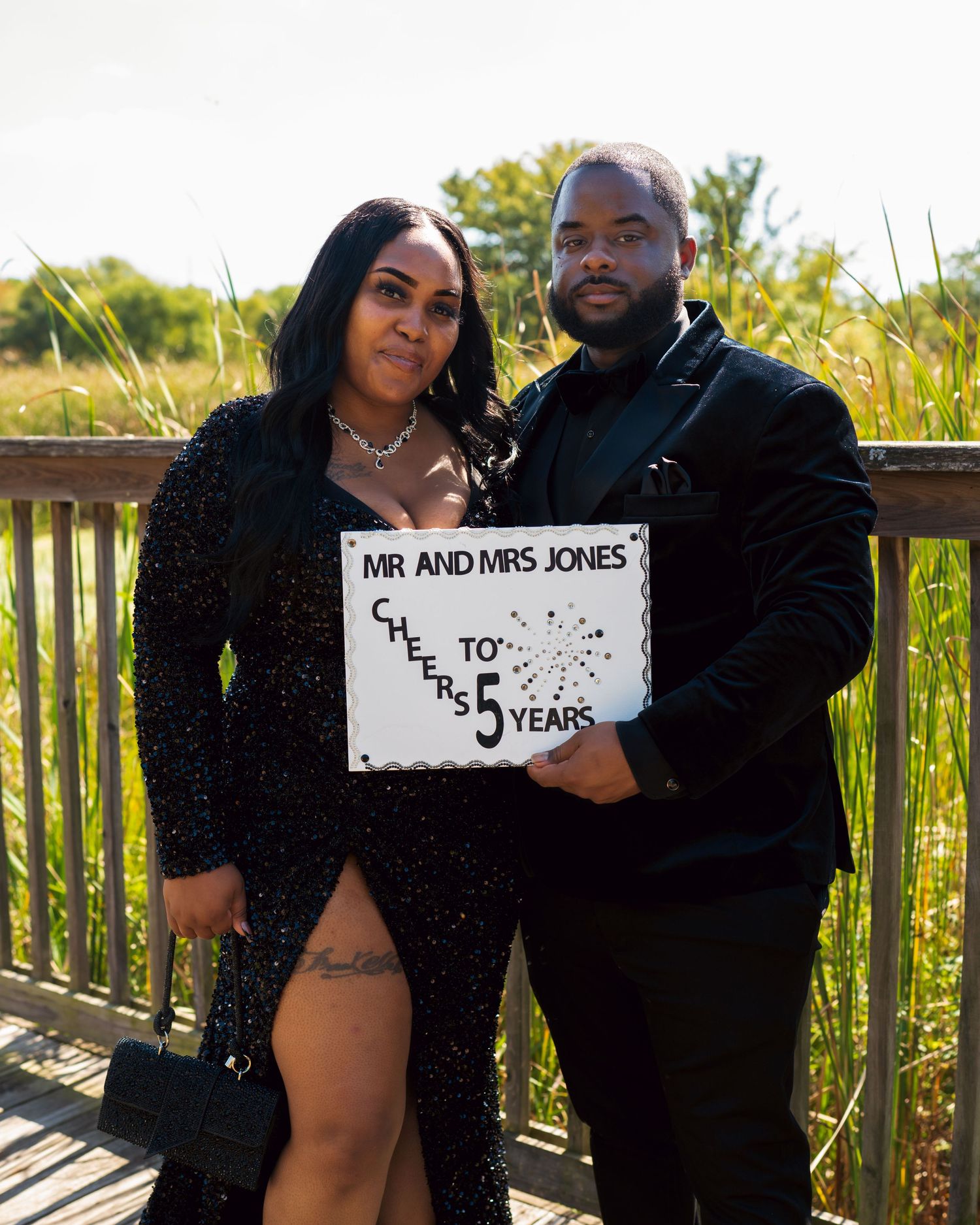 A couple poses outdoors holding a sign that reads 'Mr and Mrs Jones' while dressed in formal black attire.