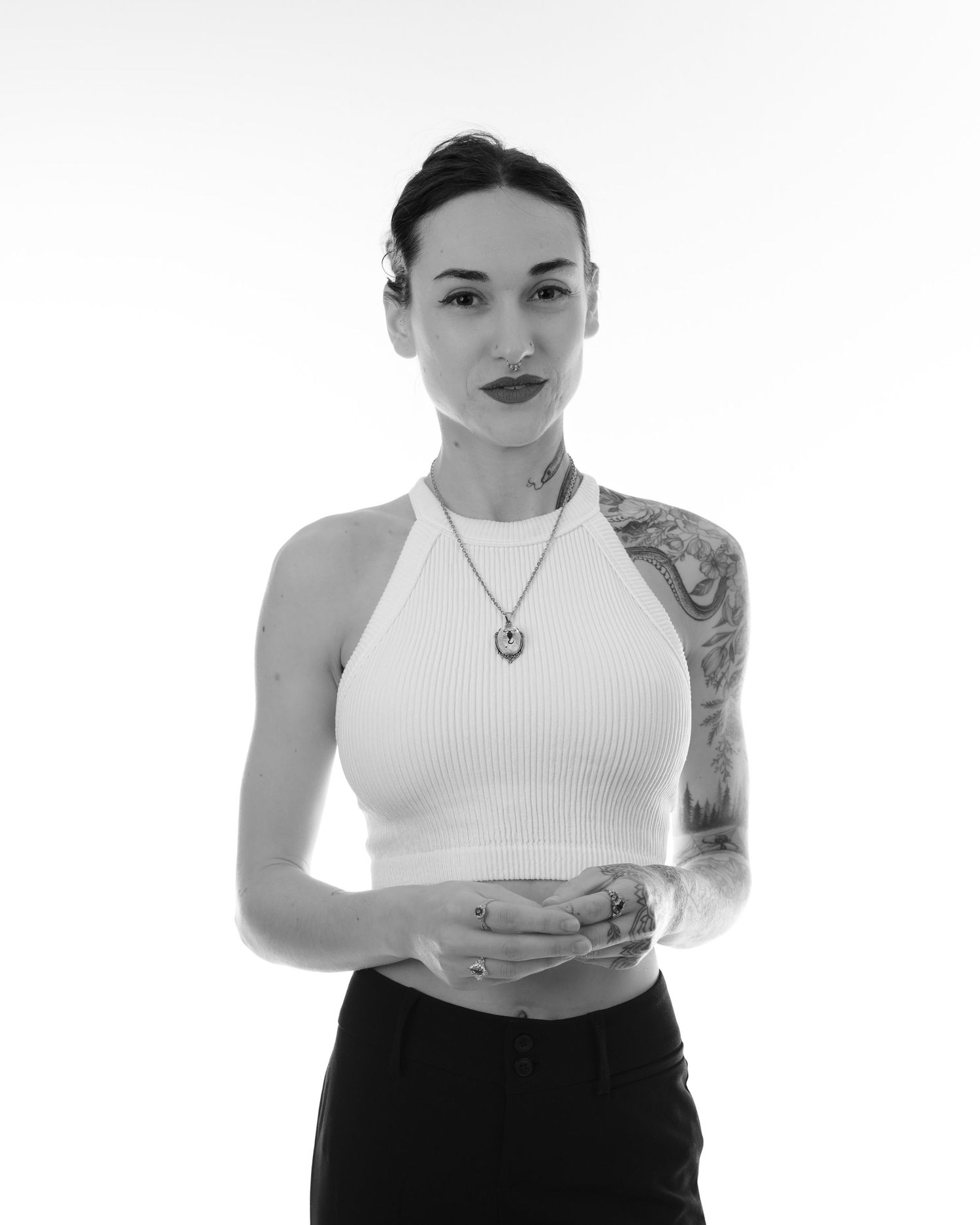 Black and white portrait of a tattooed woman in a white crop top and necklace posing against a light background.