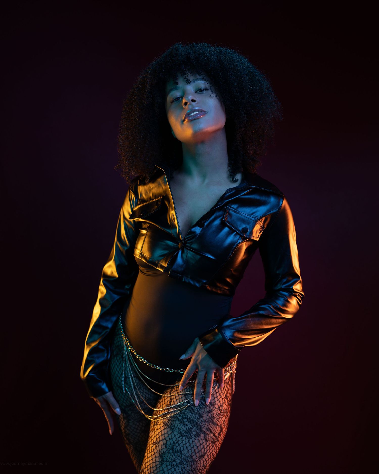 Woman in black leather jacket and chain skirt poses confidently under dramatic studio lighting with dark background.