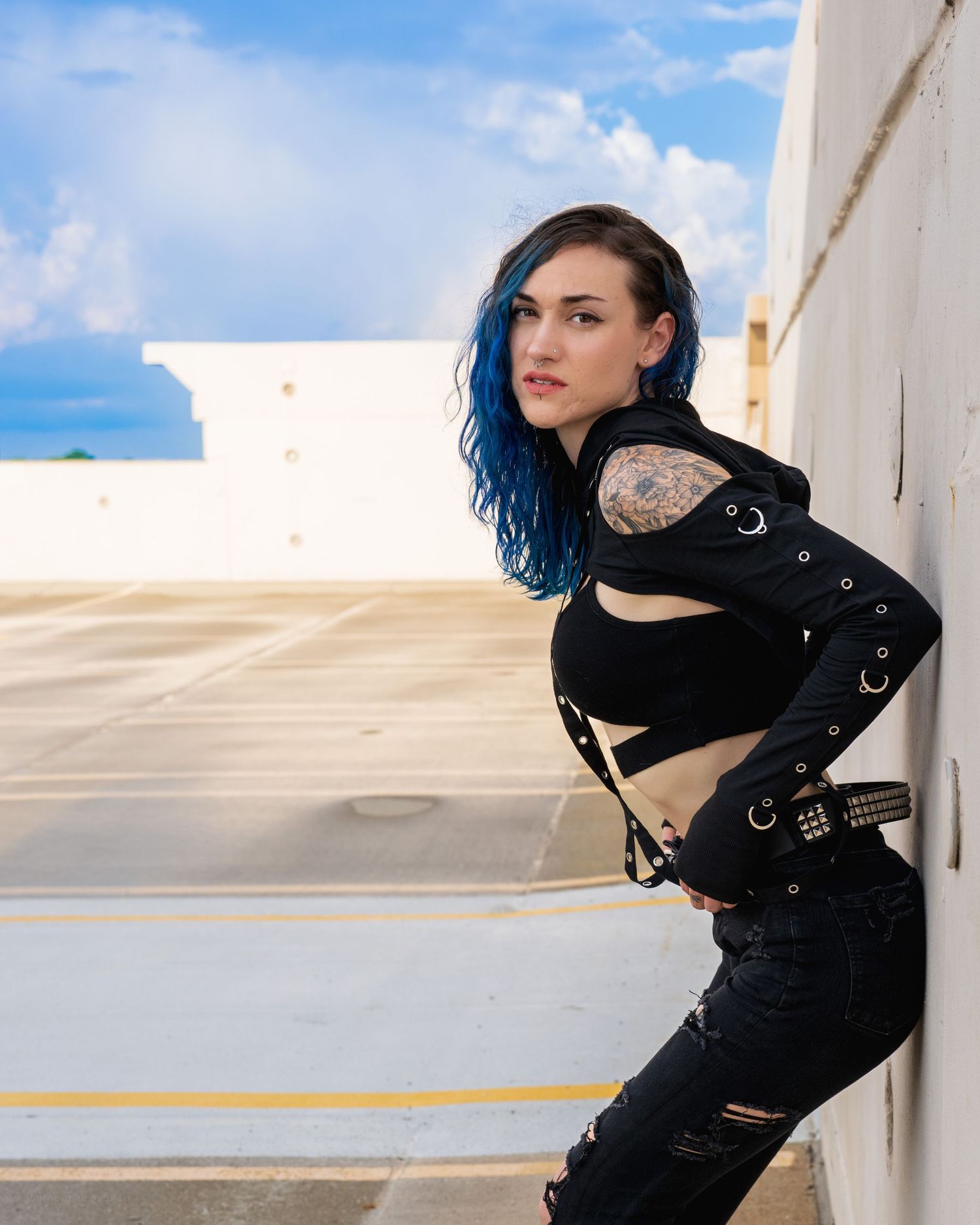 Model in black outfit with blue hair poses against white architecture and sky.
