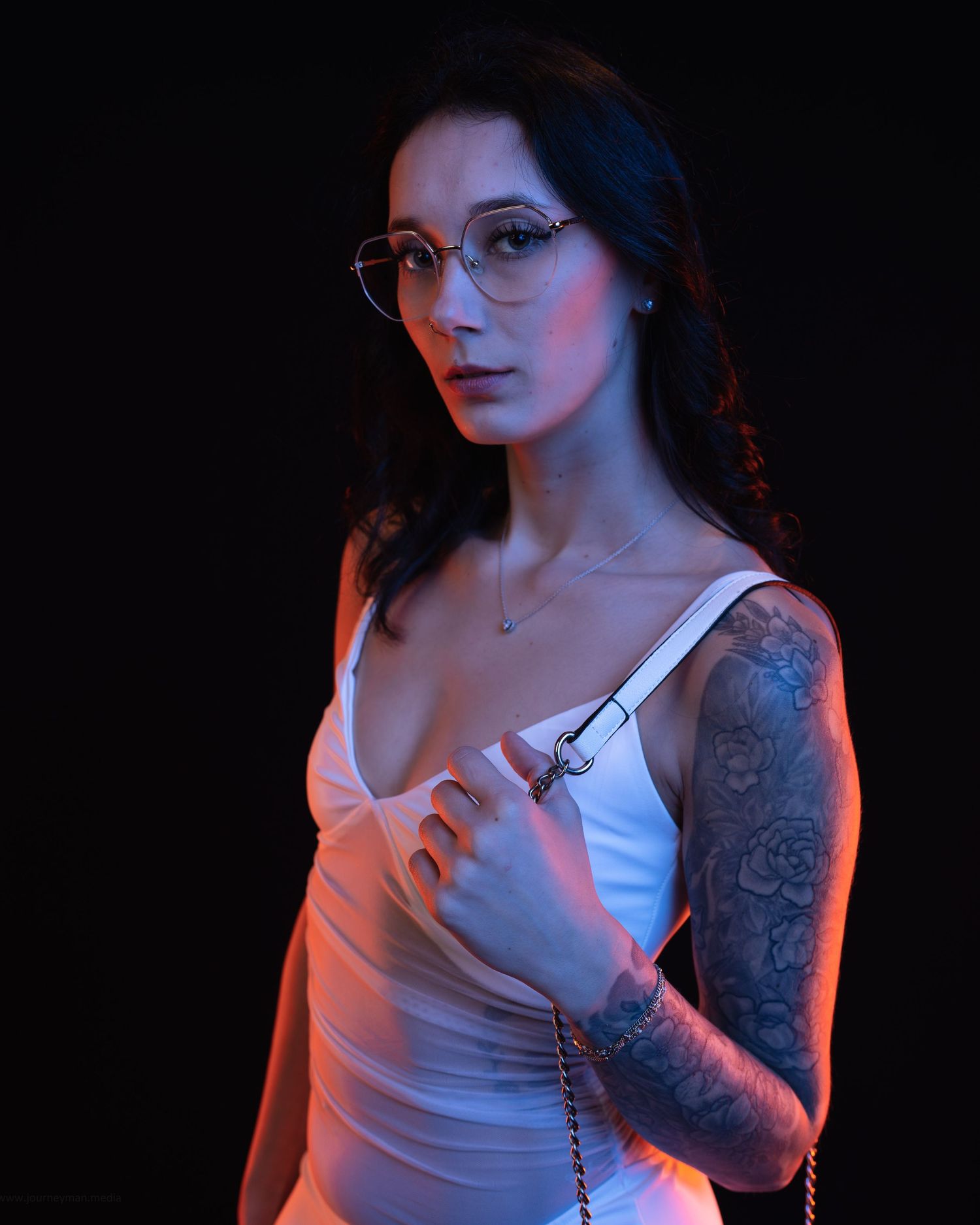 Tattooed woman in white dress and round glasses poses against dark background with blue and pink lighting.